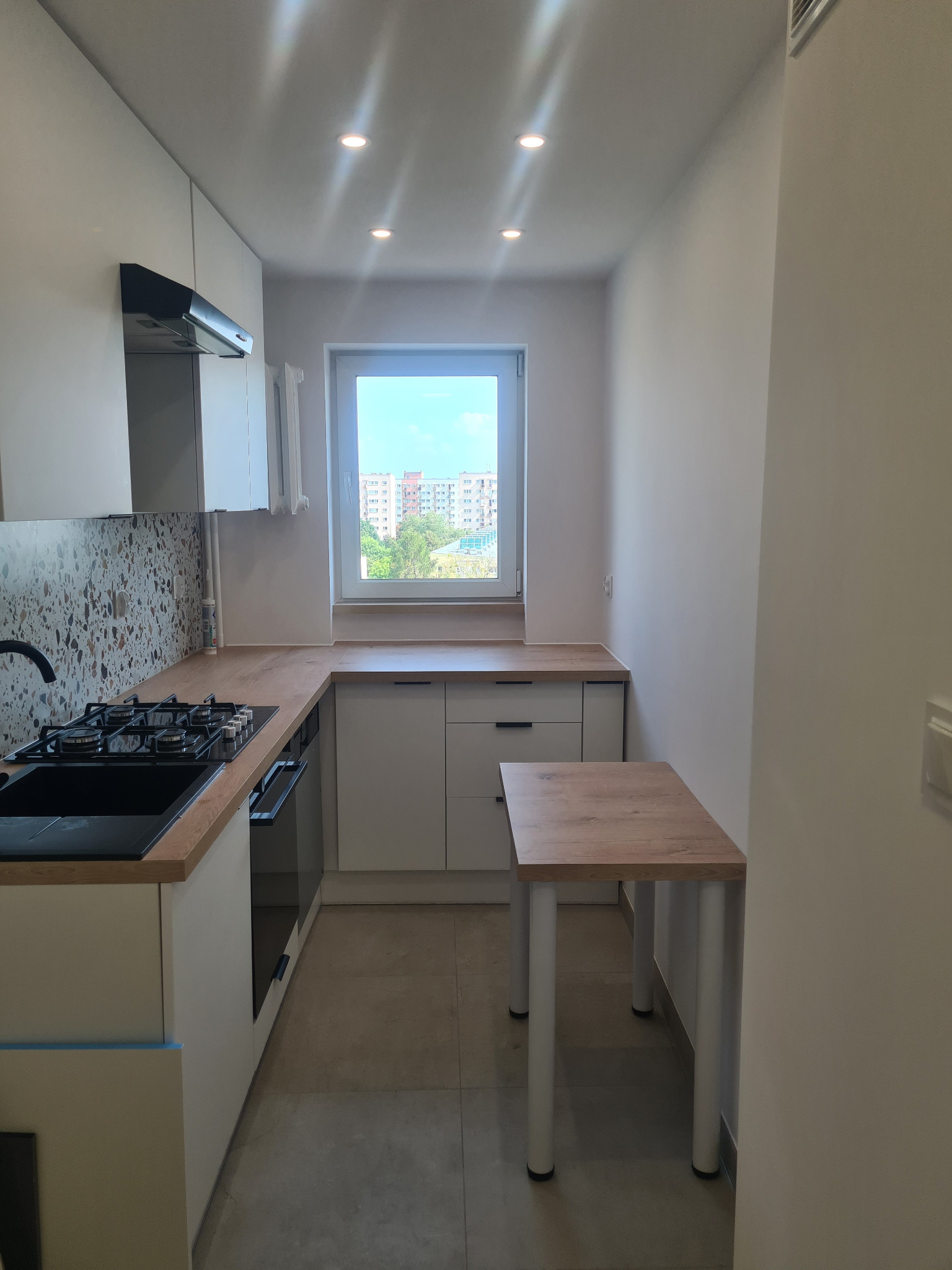 Jasna kuchnia w bloku po remoncie: białe szafki, drewniany blat, mozaika nad kuchenką, mały stół. Widok na osiedle przez okno. Minimalistyczny design.