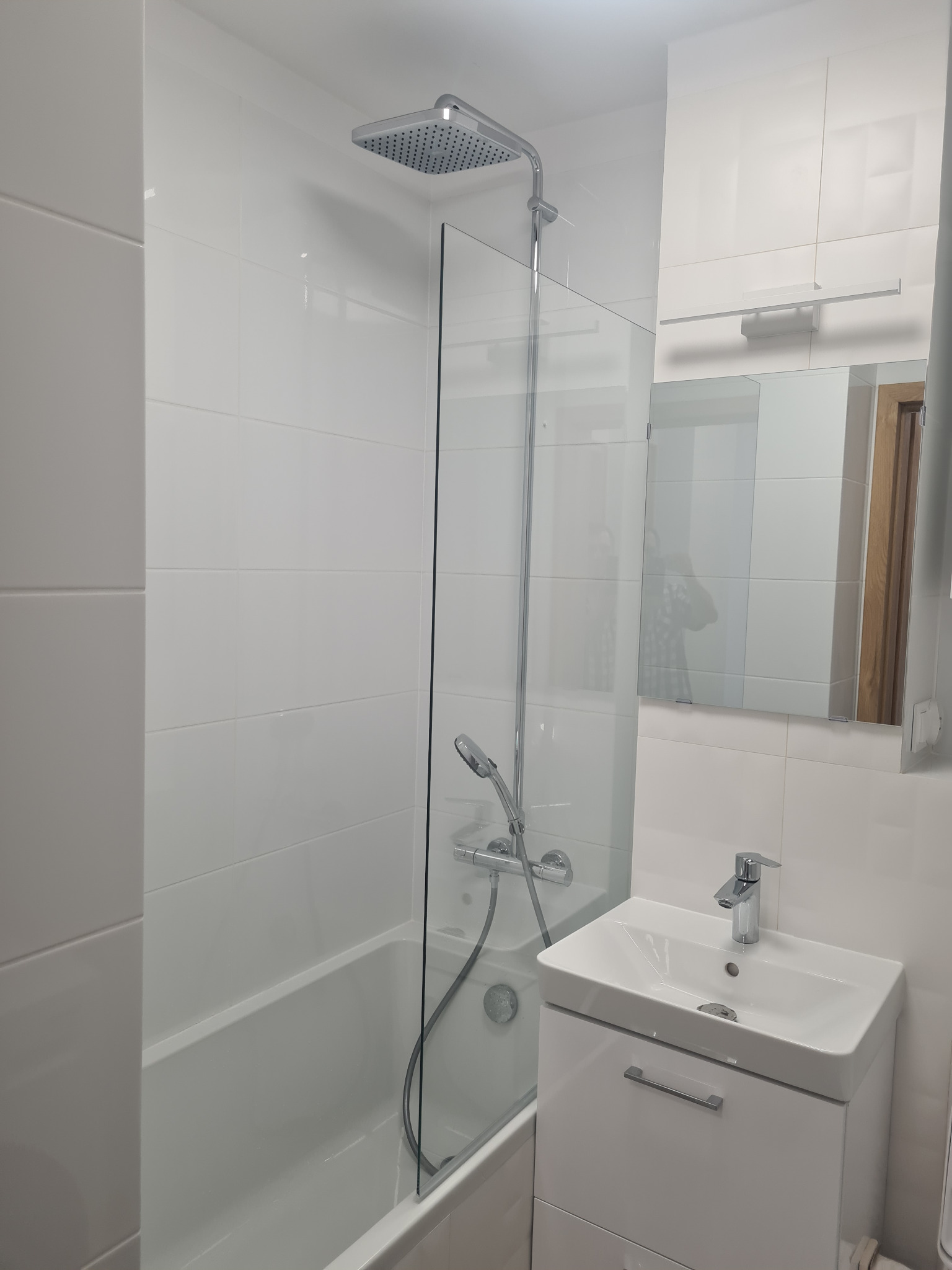 Biała łazienka po remoncie: kabina prysznicowa ze szklaną ścianką, umywalka z szafką, lustro z oświetleniem. Minimalistyczny design, jasne płytki.