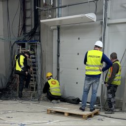 PROTOM SPÓŁKA Z OGRANICZONĄ ODPOWIEDZIALNOŚCIĄ - Trzech elektryków w kamizelkach odblaskowych z logo firmy Protrom pracuje w hali przemysłowej, instalując okablowanie elektryczne, widoczny podnośnik koszowy i brama garażowa.