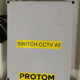 Szara skrzynka z napisem 'SWITCH CCTV #2' i 'PROTOM' na żółtych naklejkach, zamontowana na ścianie o nierównej fakturze, widoczne kable i rurki instalacyjne.