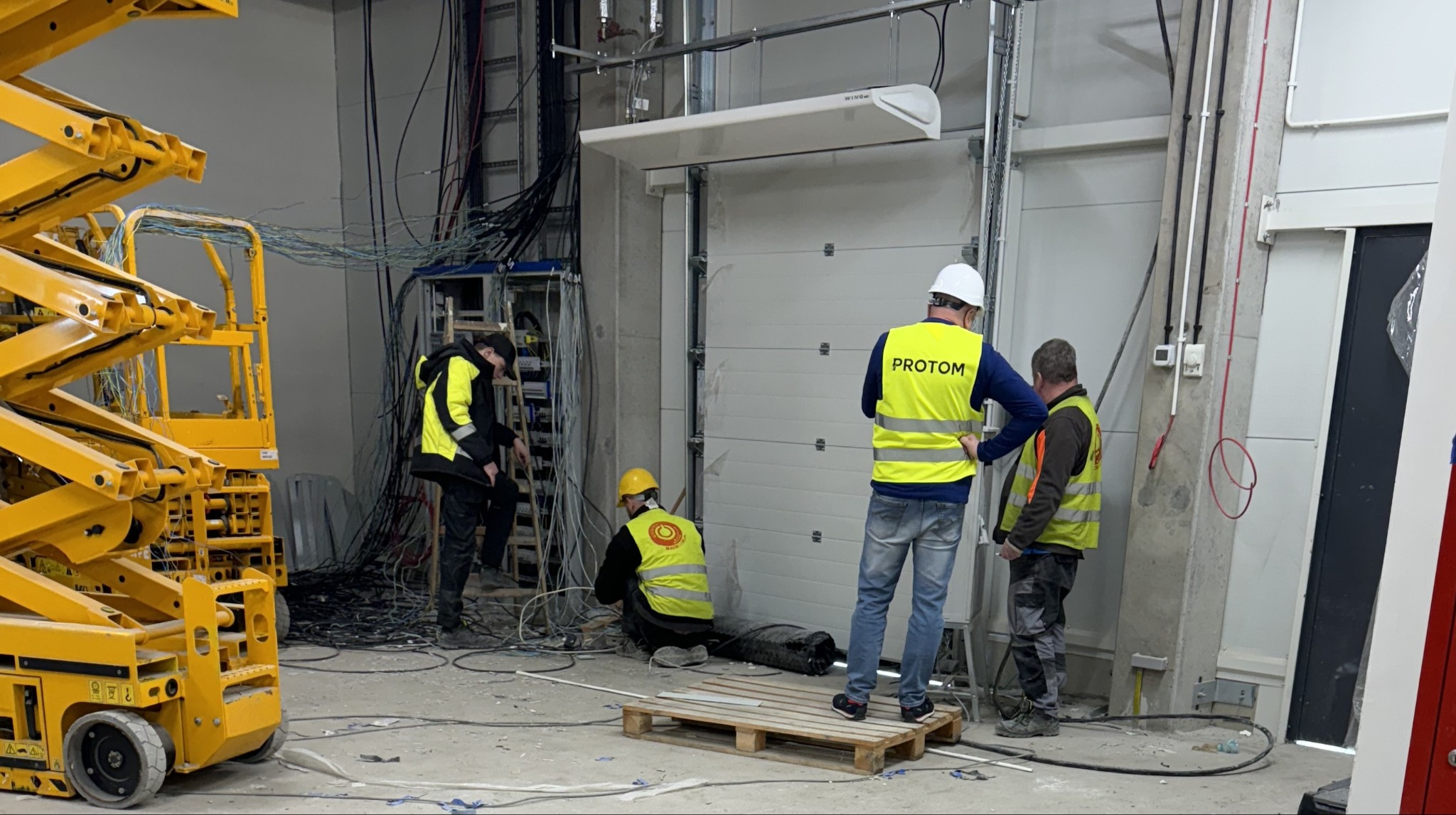 Trzech elektryków w kamizelkach odblaskowych z logo firmy Protrom pracuje w hali przemysłowej, instalując okablowanie elektryczne, widoczny podnośnik koszowy i brama garażowa.