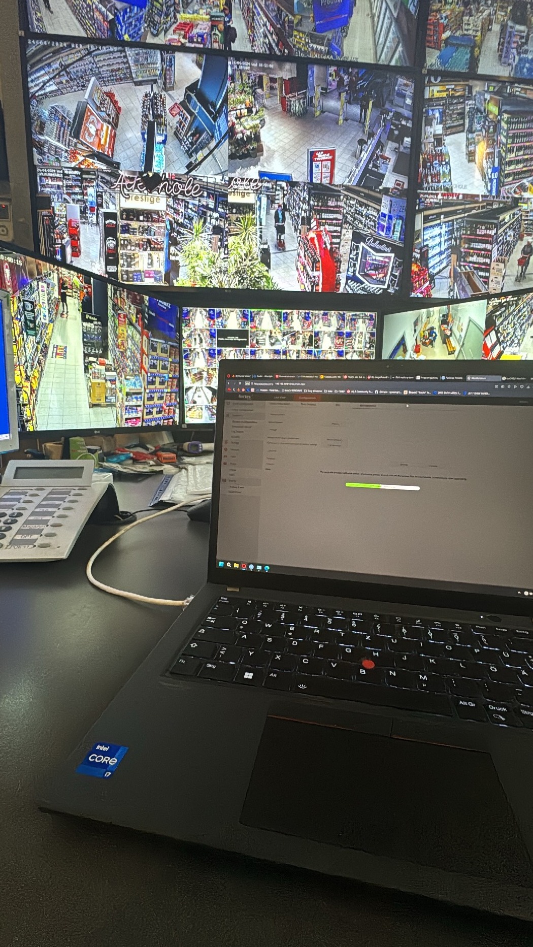 Ekran laptopa z widocznym paskiem postępu ładowania, w tle monitory z podglądem z kamer monitoringu w supermarkecie, obok telefon stacjonarny.