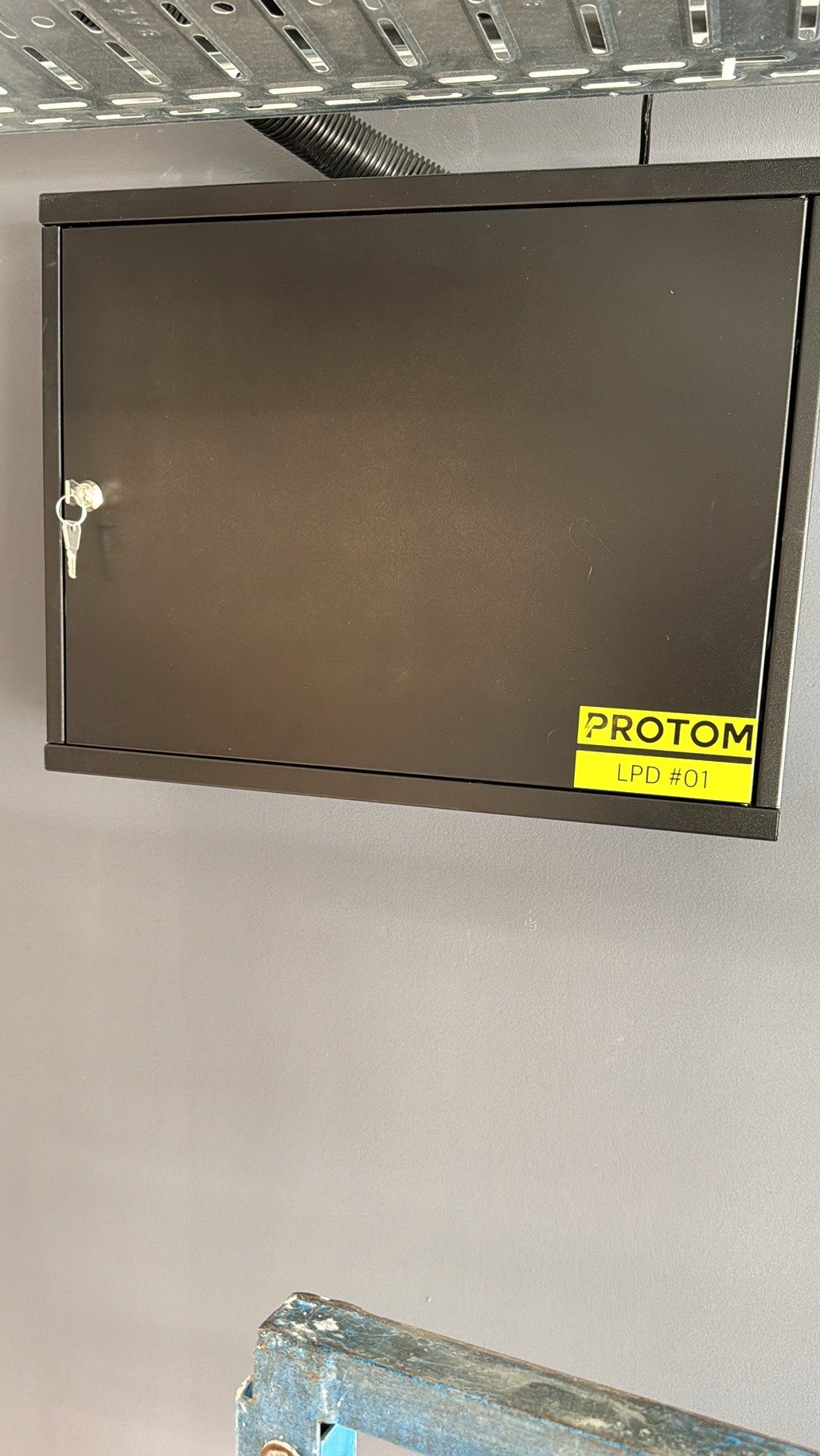 Zamknięta, metalowa szafka elektryczna w kolorze ciemnoszarym, zamontowana na szarej ścianie, z kluczykiem w zamku i żółtą naklejką 'PROTOM LPD #01'. Nad szafką widoczna metalowa drabinka kablowa...