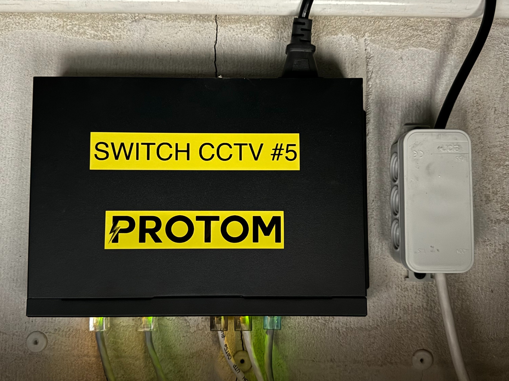 Czarna skrzynka SWITCH CCTV #5 PROTOM z podłączonymi przewodami sieciowymi i kablem zasilającym, zamontowana na szarej ścianie obok puszki elektrycznej.