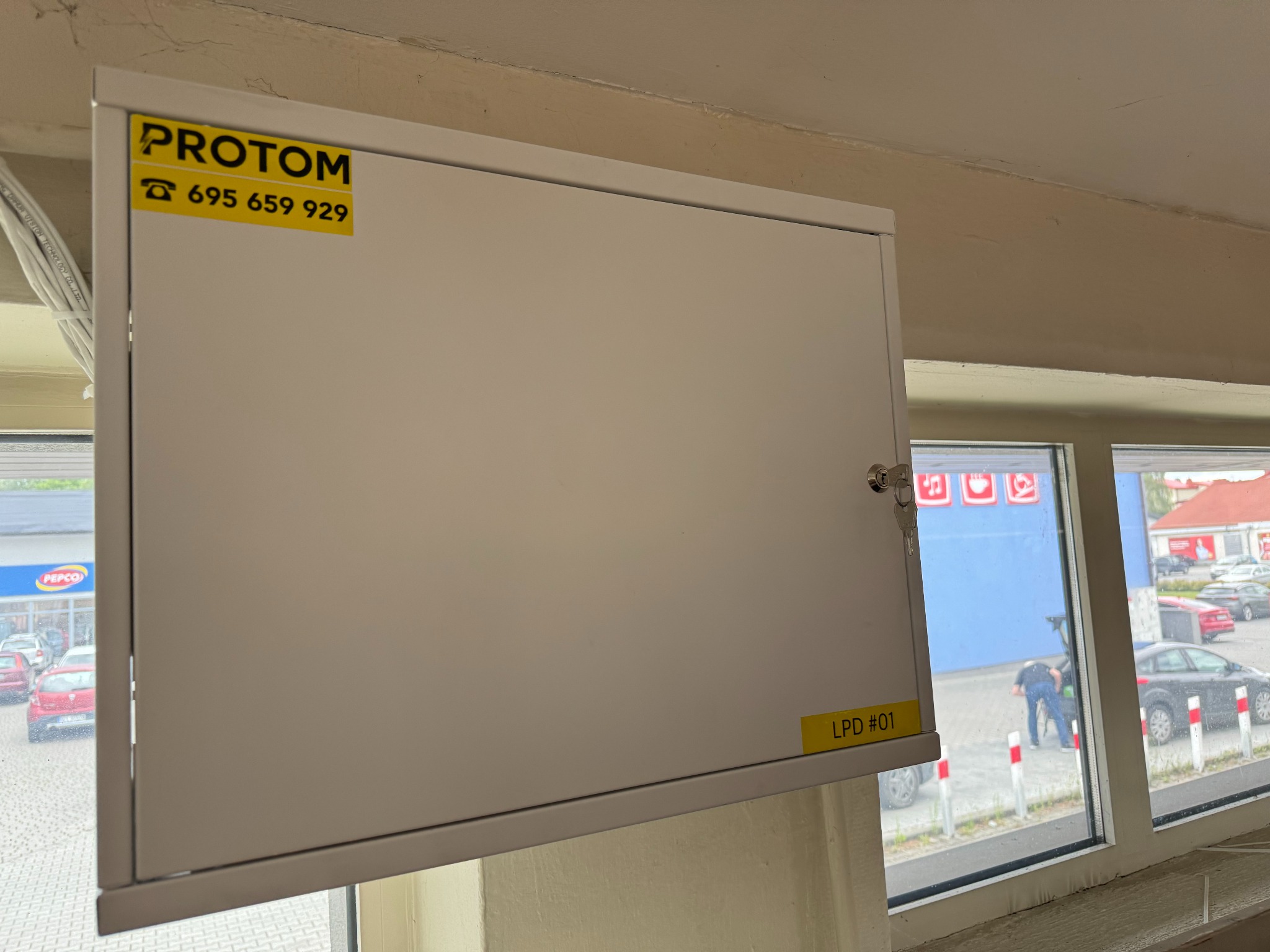 Biała skrzynka elektryczna z naklejką firmy 'PROTOM' i numerem telefonu, zamontowana w pomieszczeniu z widokiem na parking sklepu Pepco.