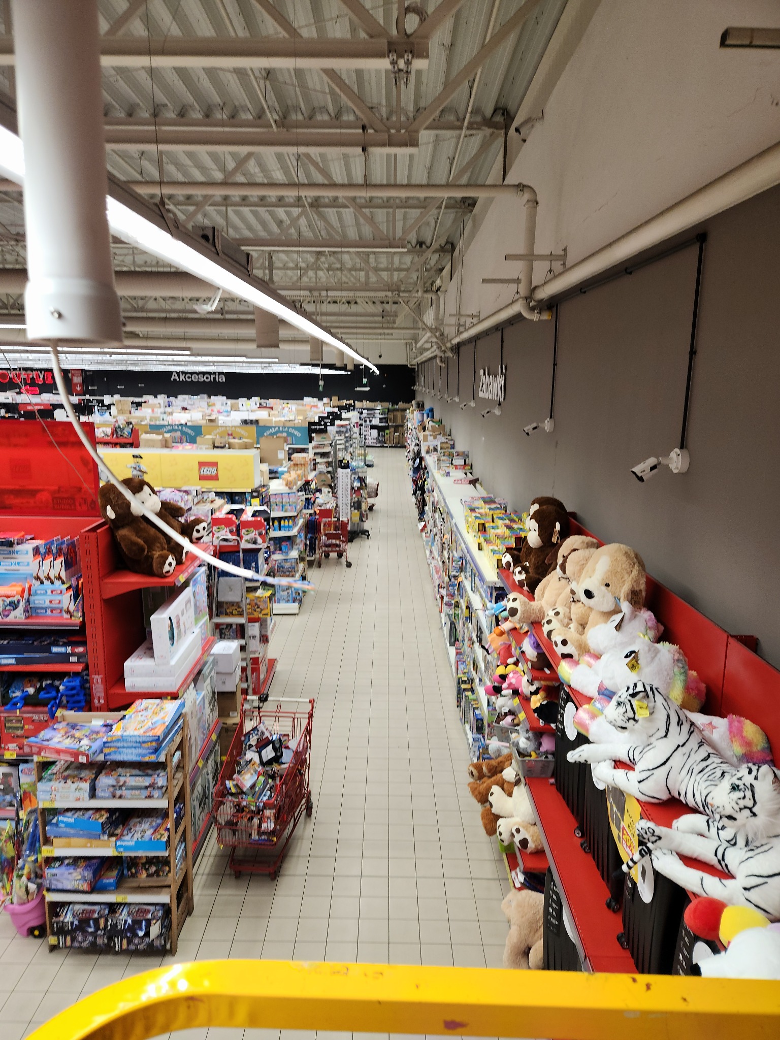 Długi korytarz sklepu z zabawkami, wypełniony regałami z pluszakami, grami i innymi artykułami dla dzieci, oświetlony przez rzędy lamp sufitowych i monitorowany przez kamery przemysłowe.
