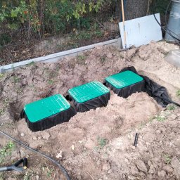 GREEN Eden - Trzy zielone pokrywy skrzynek zaworowych w ziemi, prawdopodobnie część instalacji nawadniającej, widoczne czarne rury i folia ochronna, ziemia wokół wykopana, w tle ogrodzenie i roślinność.