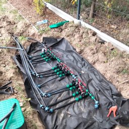 GREEN Eden - Fragment instalacji nawadniającej z czarnymi rurami, zielonymi złączkami i czerwonymi przewodami elektrycznymi, rozłożony na czarnej folii w wykopie ziemnym, widoczne narzędzia i skrzynka.