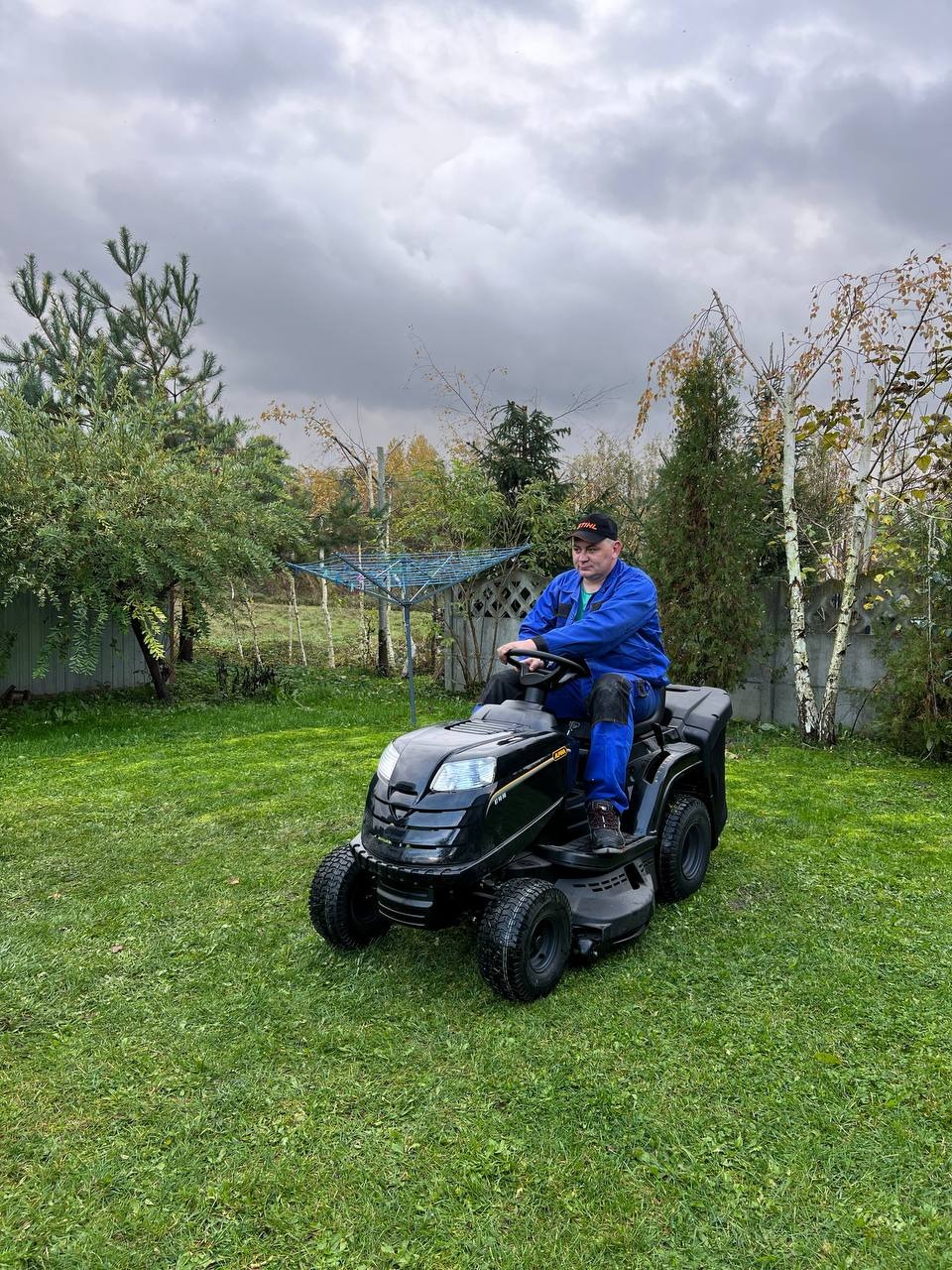 Mężczyzna w niebieskim kombinezonie i czapce na czarnej kosiarce traktorowej na tle zielonego trawnika i pochmurnego nieba.
