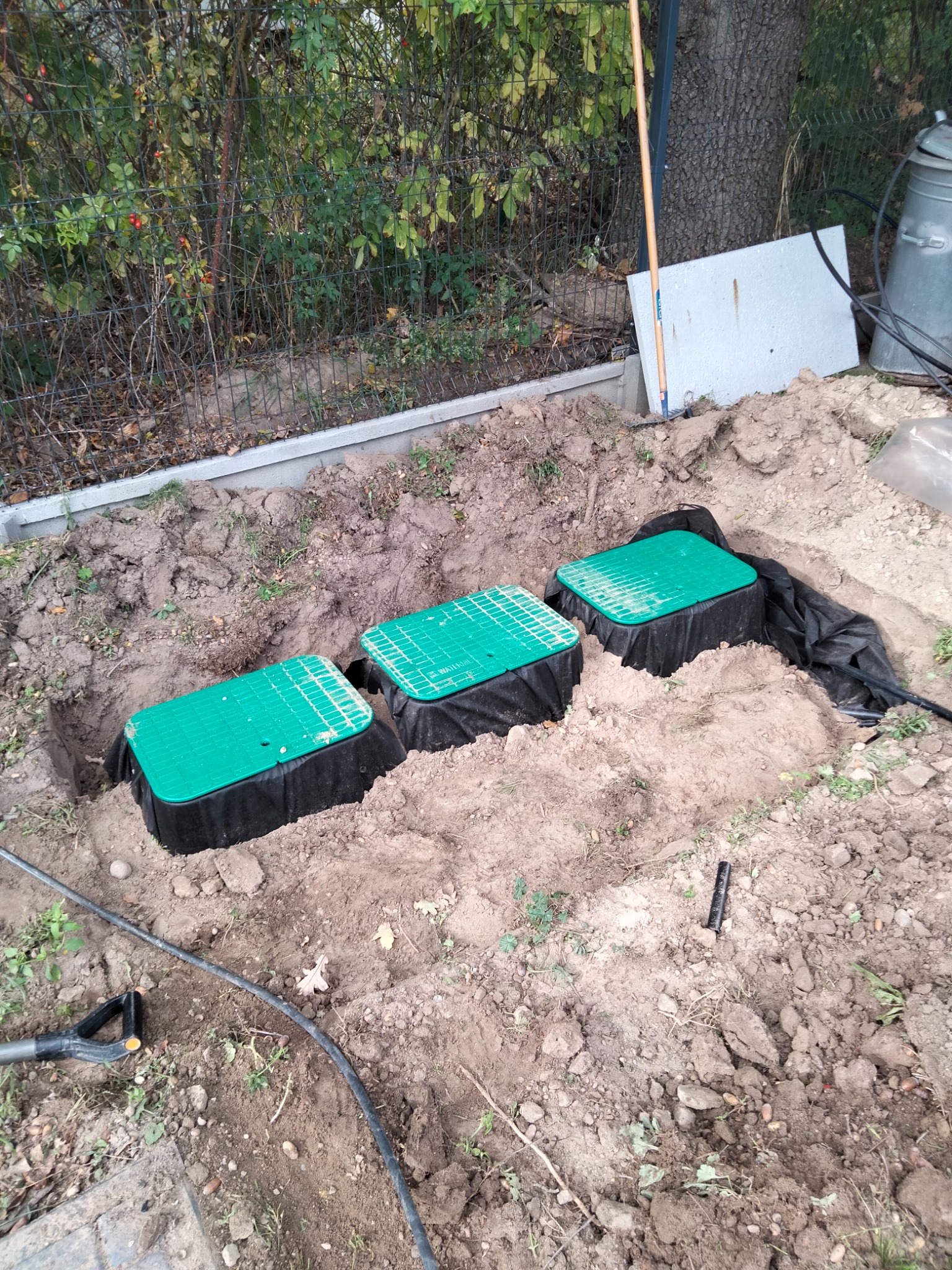 Trzy zielone pokrywy skrzynek zaworowych w ziemi, prawdopodobnie część instalacji nawadniającej, widoczne czarne rury i folia ochronna, ziemia wokół wykopana, w tle ogrodzenie i roślinność.