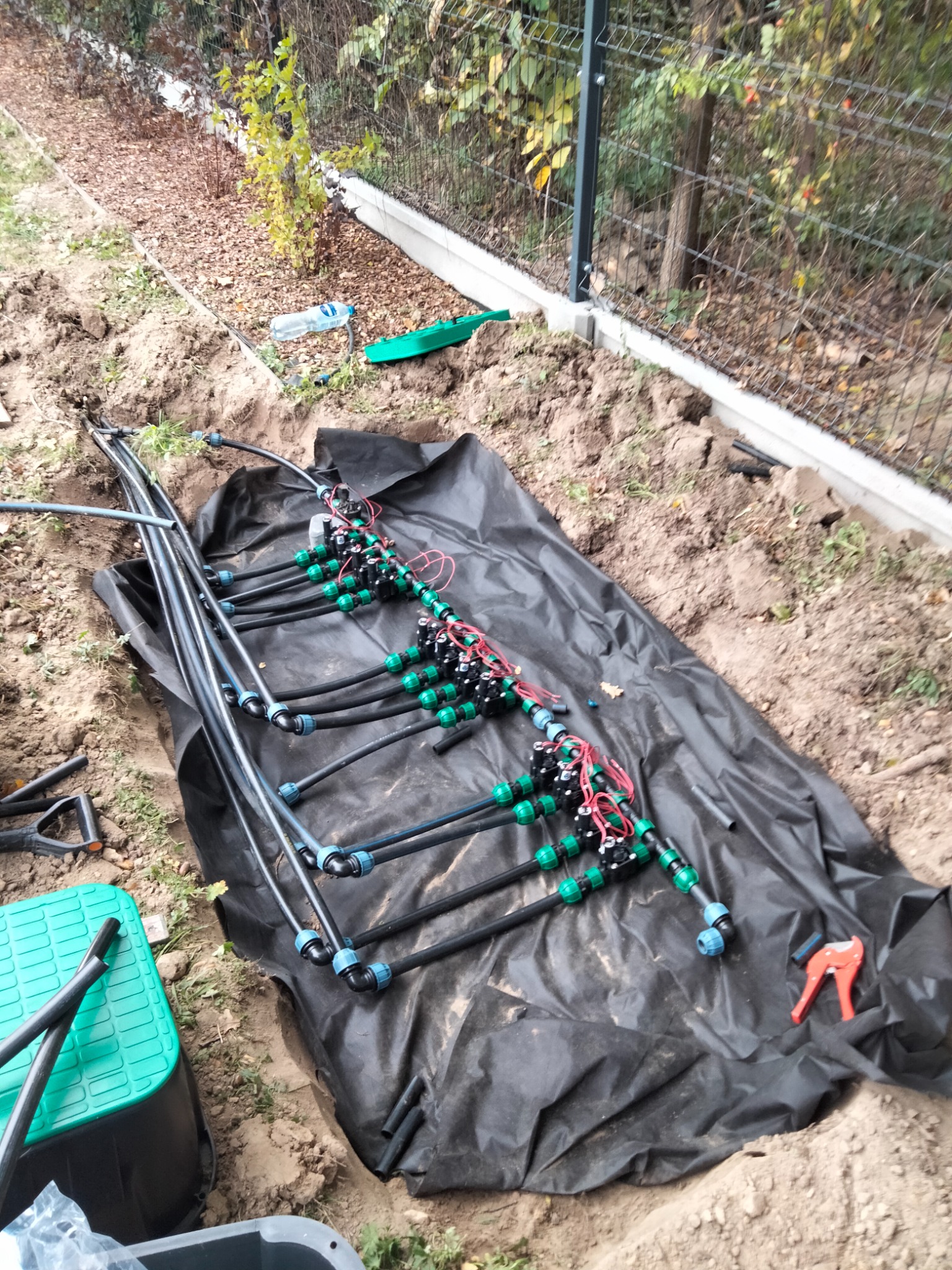Fragment instalacji nawadniającej z czarnymi rurami, zielonymi złączkami i czerwonymi przewodami elektrycznymi, rozłożony na czarnej folii w wykopie ziemnym, widoczne narzędzia i skrzynka.