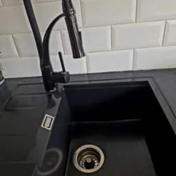 Czarny zlewozmywak granitowy z czarną baterią na tle białych, prostokątnych płytek.