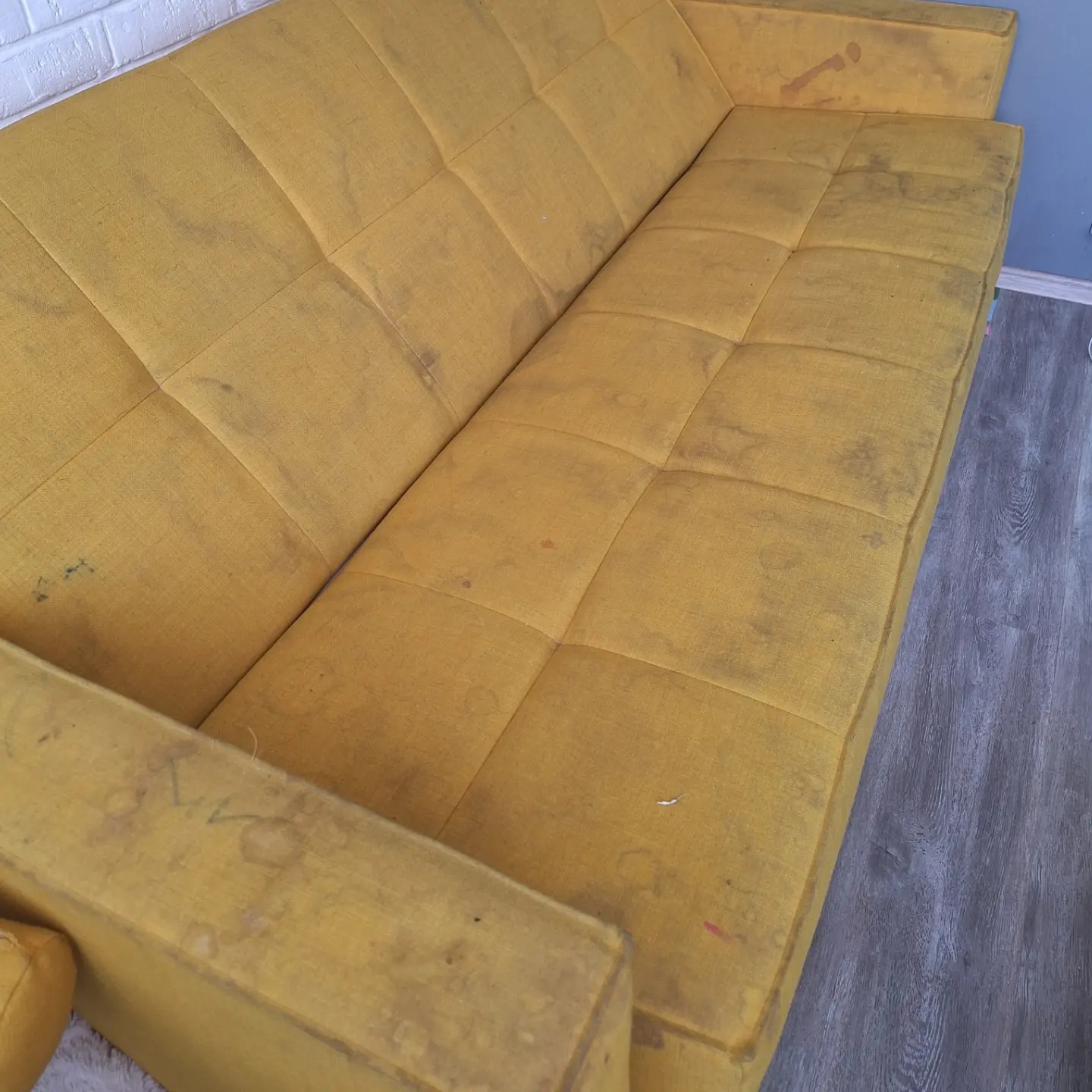 Żółta, brudna kanapa z widocznymi plamami i zabrudzeniami na tapicerce, sfotografowana z góry na tle ściany z białej cegły i szarej podłogi imitującej drewno.