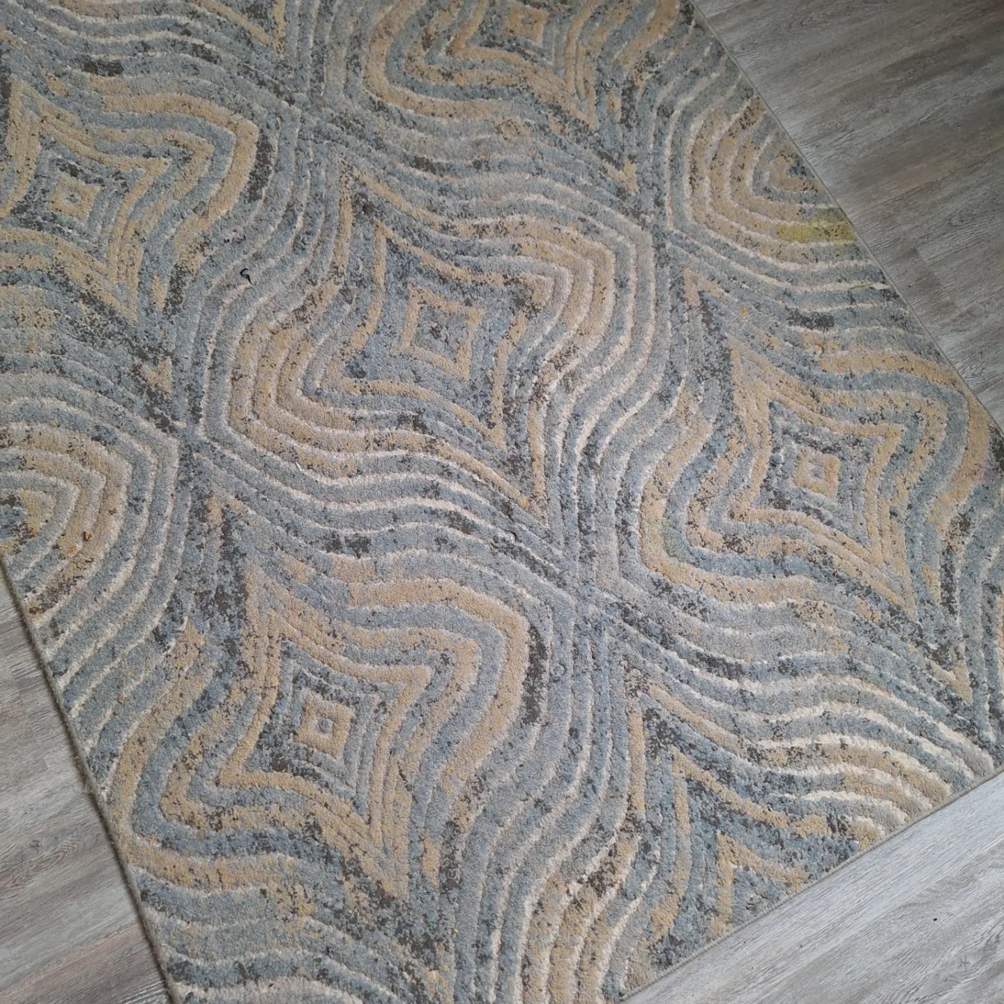 Beżowo-szary dywan z wzorem przypominającym warstwy geologiczne, leżący na szarych panelach podłogowych.