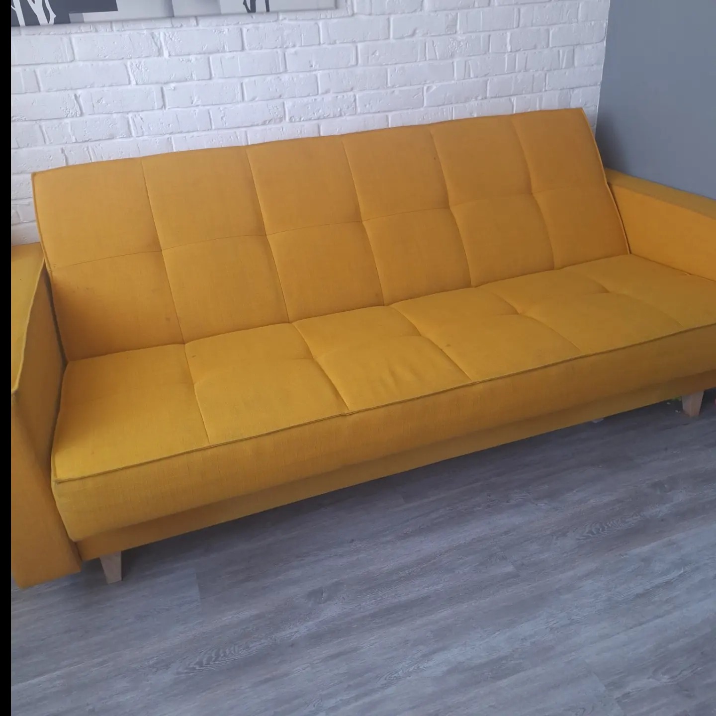 Żółta kanapa z tkaniny z wyraźnymi przeszyciami na tle białej ściany z cegły i szarych paneli podłogowych.