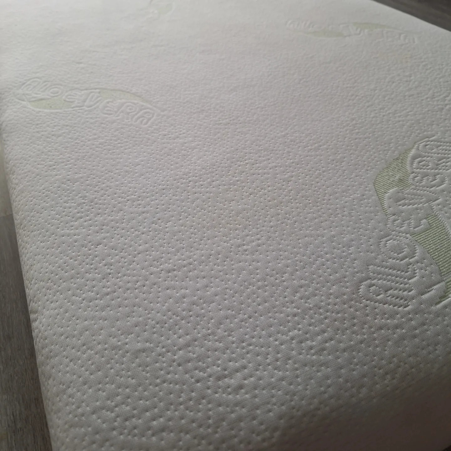 Jasny materac z tłoczonym wzorem i logo 'Aloe Vera', leżący na podłodze z paneli.