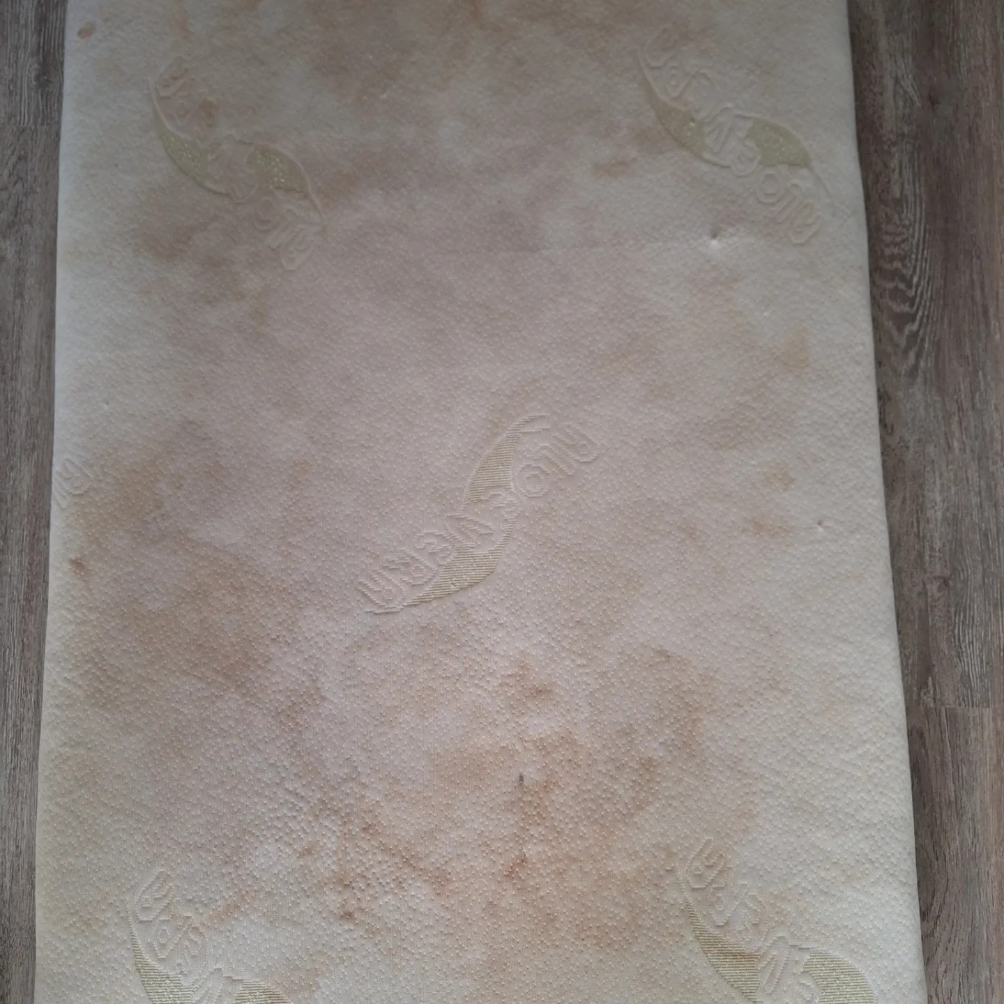 Widok z góry na mocno zabrudzony materac z widocznym wzorem i plamami, leżący częściowo na podłodze imitującej drewno.