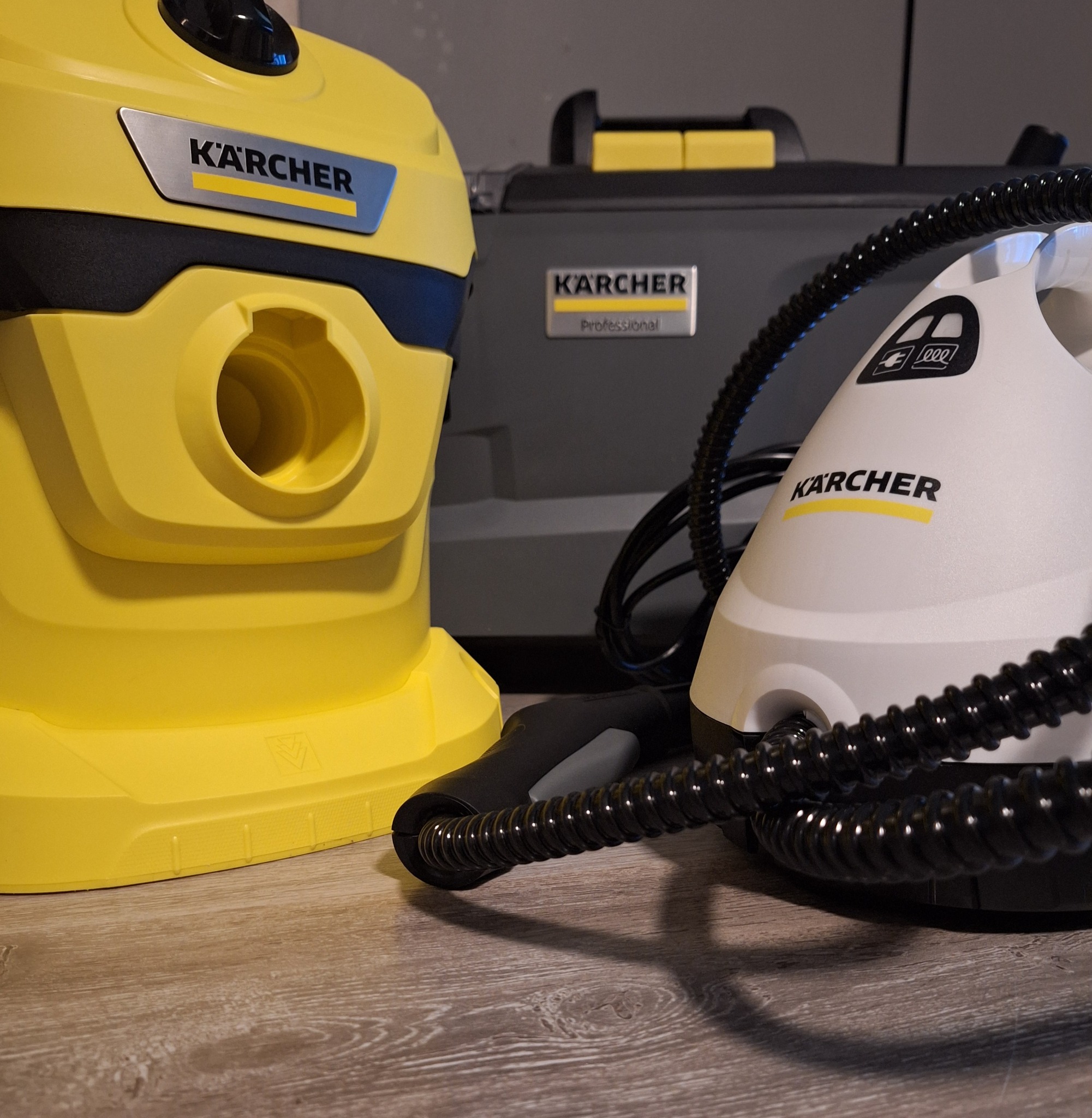 Żółty odkurzacz Karcher, parownica Karcher z czarnym wężem i szare pudełko Karcher Professional na drewnianej podłodze.