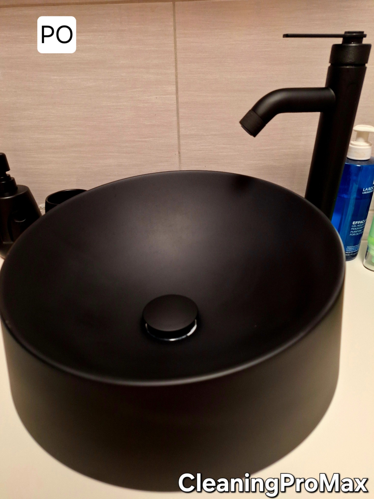 Czarna, okrągła umywalka nablatowa z czarną armaturą i kosmetykami w tle, widok z góry.