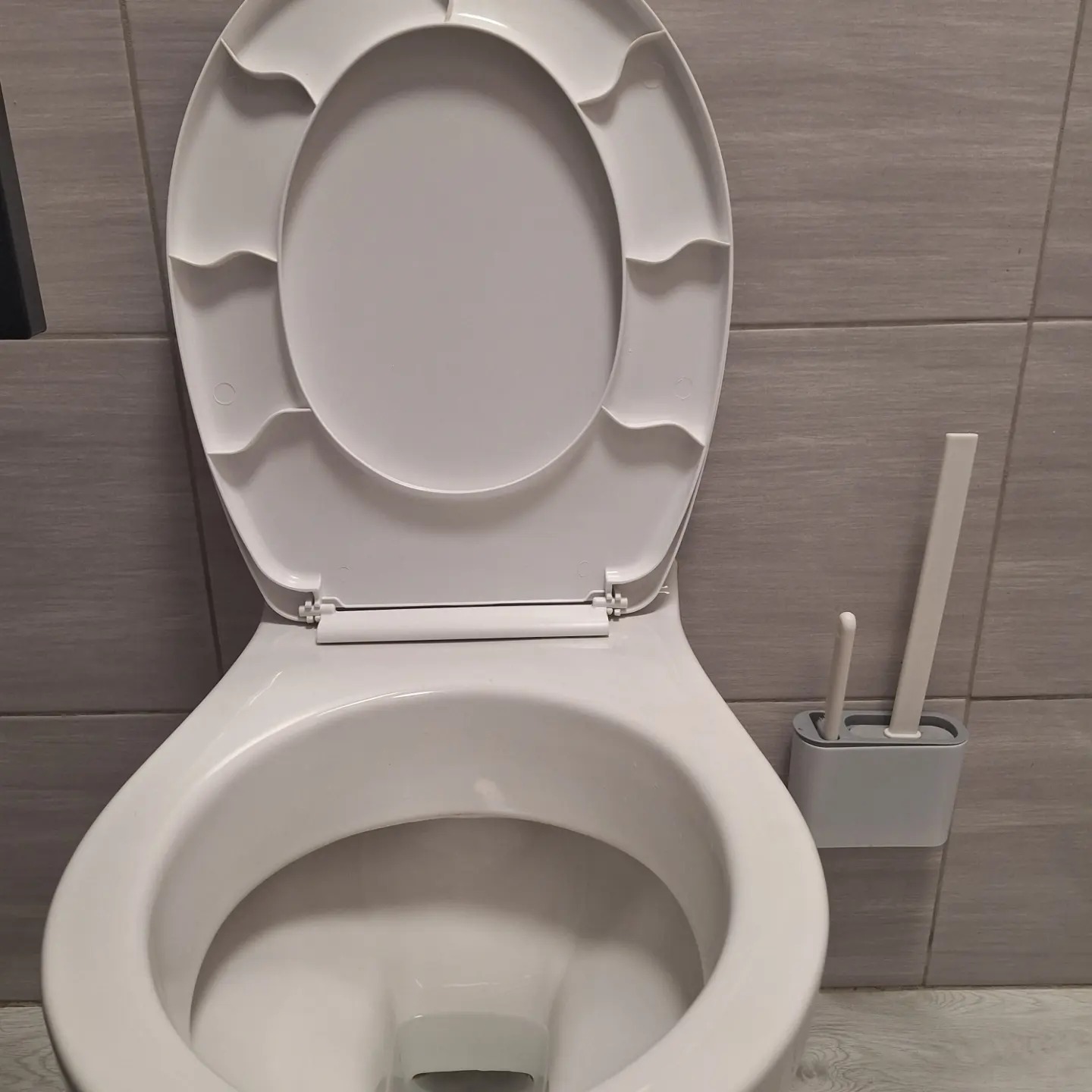 Otwarta, biała toaleta z ceramiczną muszlą i deską, widoczna szczotka do WC w szarym pojemniku na ścianie z płytek.