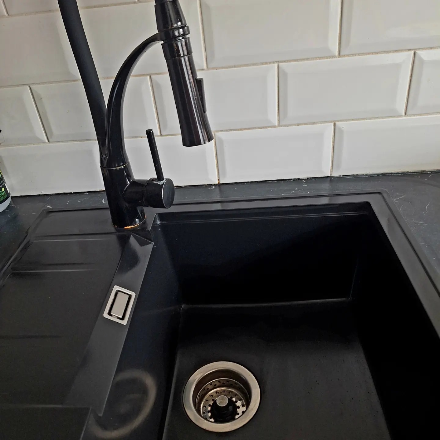 Czarny zlewozmywak granitowy z czarną baterią na tle białych, prostokątnych płytek.