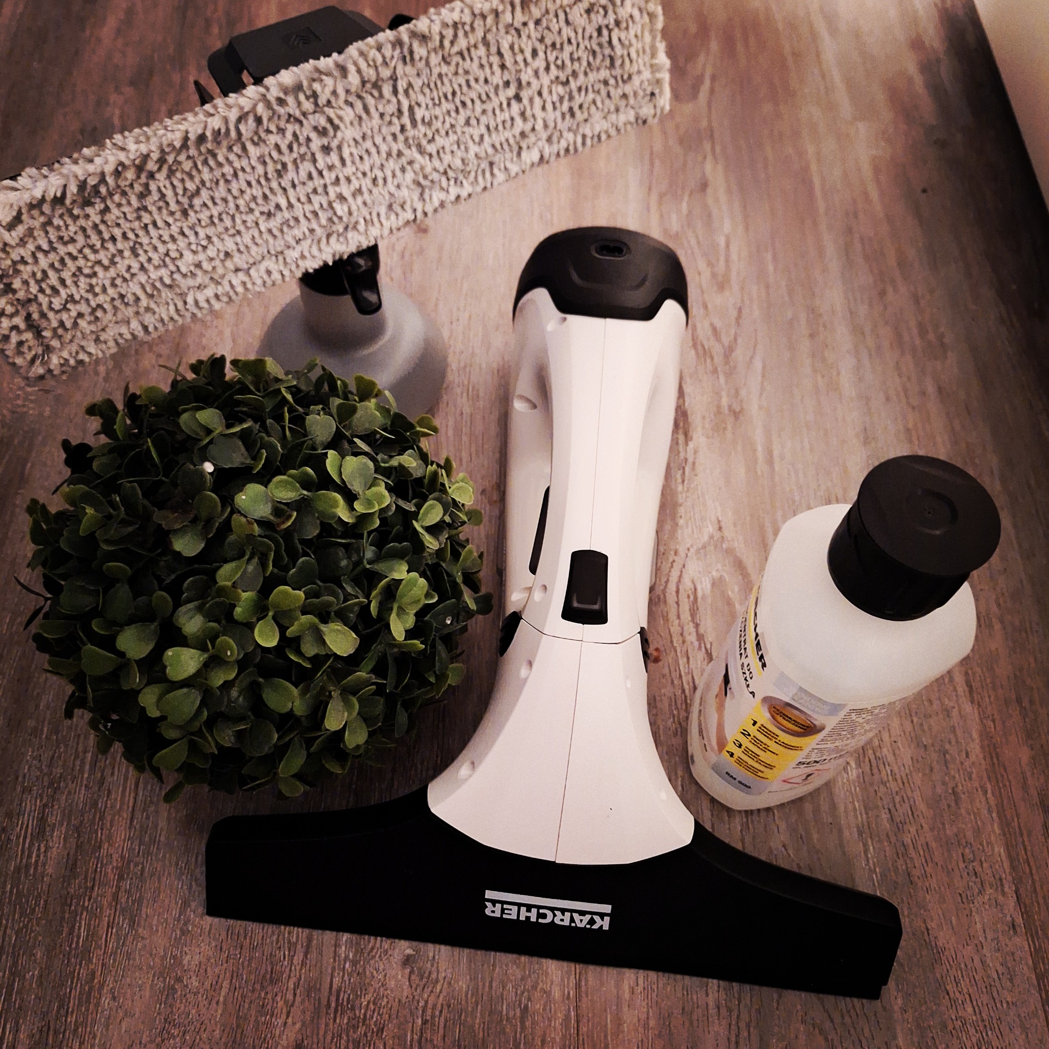 Zestaw do mycia okien Kärcher z płynem i nakładką z mikrofibry, obok dekoracyjna kula z zielonych listków, ułożony na drewnopodobnej powierzchni.