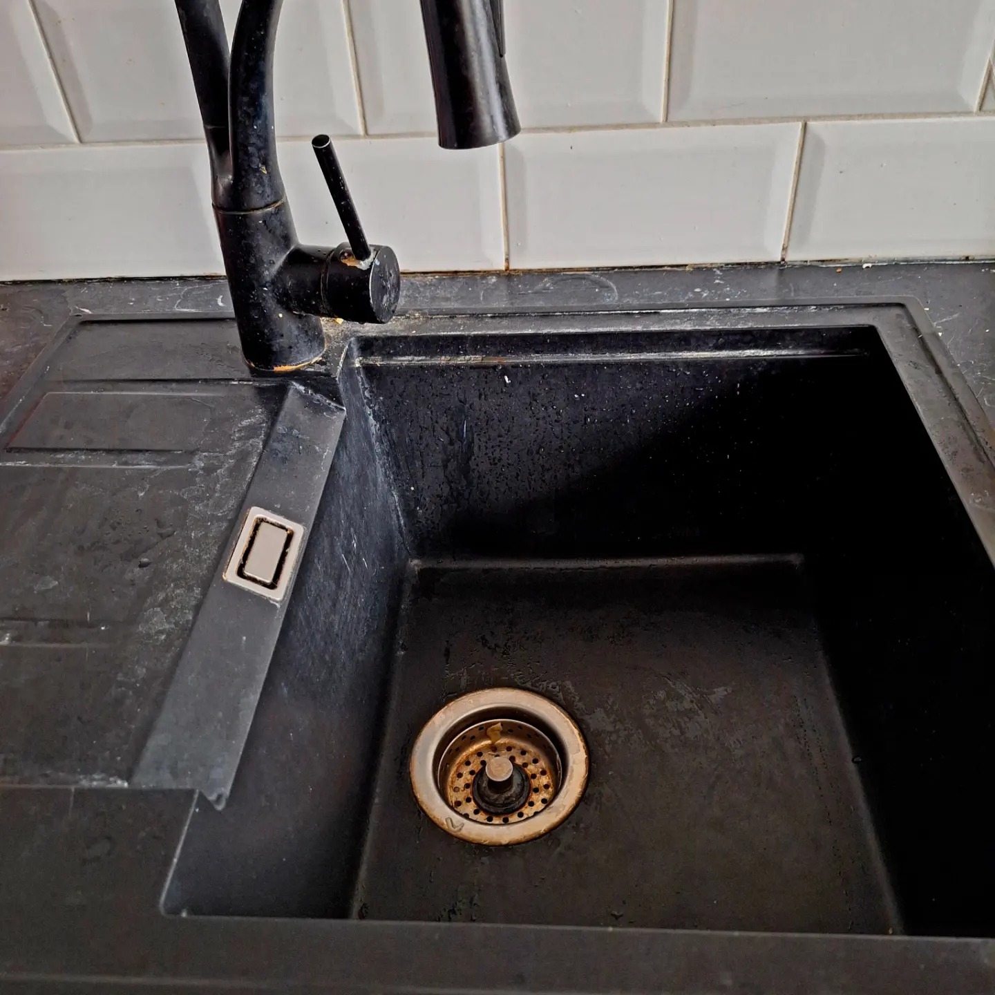 Czarny, zabrudzony zlew kuchenny z baterią, widoczne osady i plamy na powierzchni, białe płytki w tle.