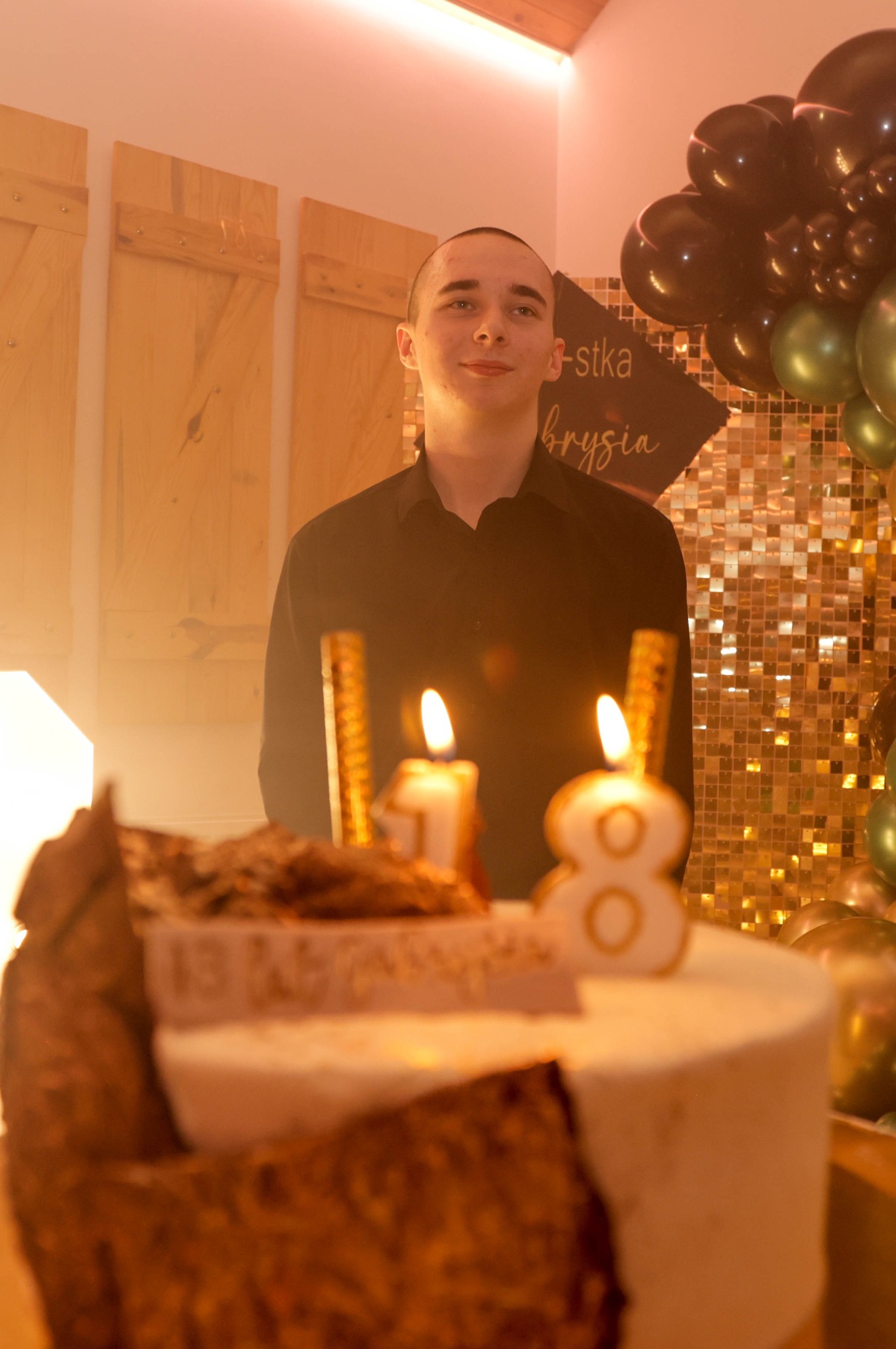 Młody mężczyzna w Miechowie uśmiecha się za tortem urodzinowym z płonącymi świeczkami w kształcie cyfr 1 i 8, w tle złota ściana cekinowa i balony.