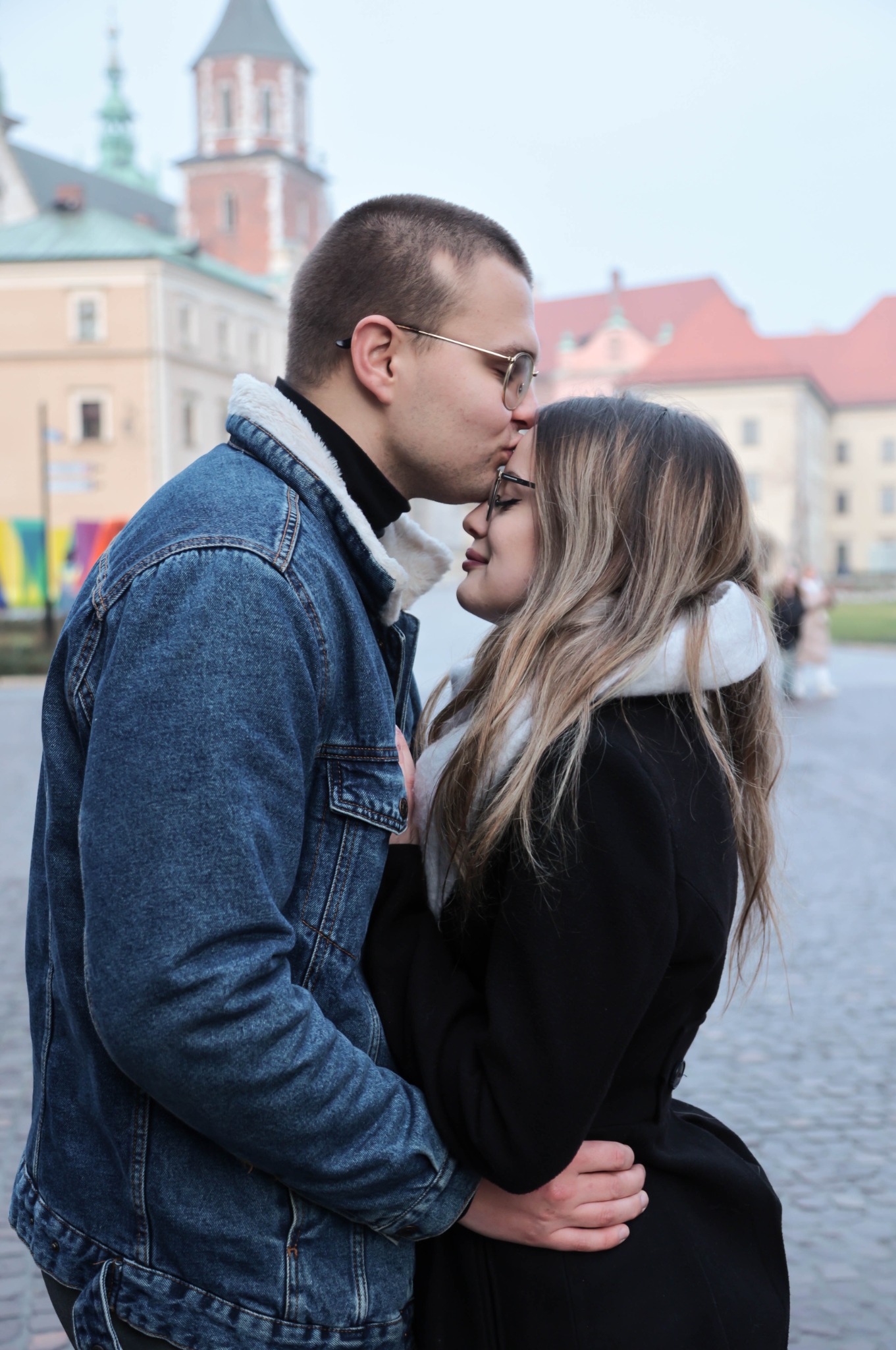 Młoda para w objęciach na tle Wawelu: mężczyzna w dżinsowej kurtce z kożuszkiem całuje kobietę w czoło, oboje w okularach.