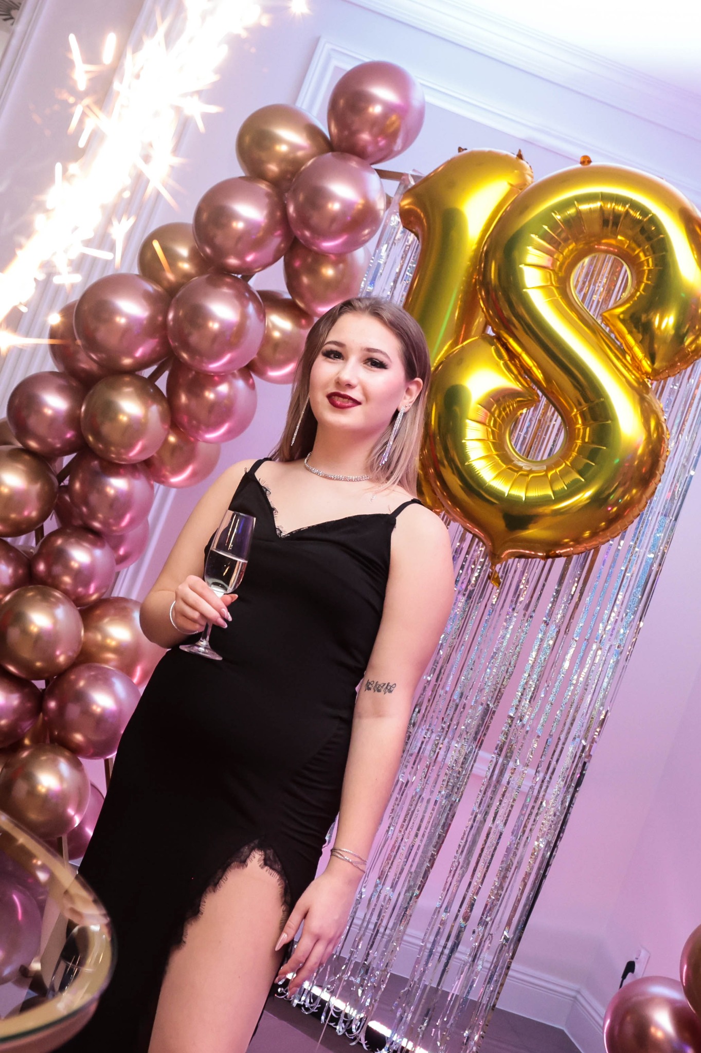 Młoda kobieta w czarnej sukience pozuje z kieliszkiem szampana na tle dekoracji z różowych balonów, fontanny iskier i złotych balonów w kształcie cyfry 18.