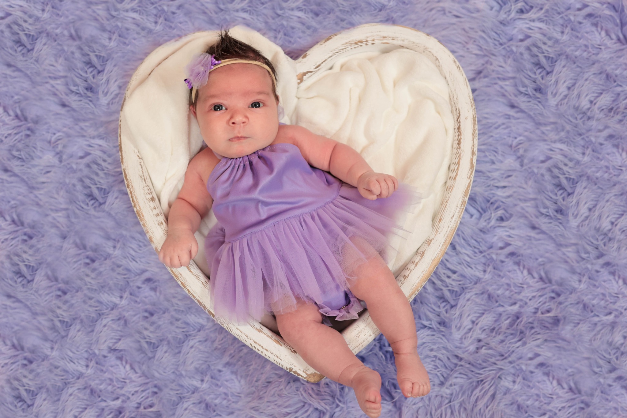 Noworodek w fioletowej sukience leży w drewnianym koszu w kształcie serca na puszystym, fioletowym tle.