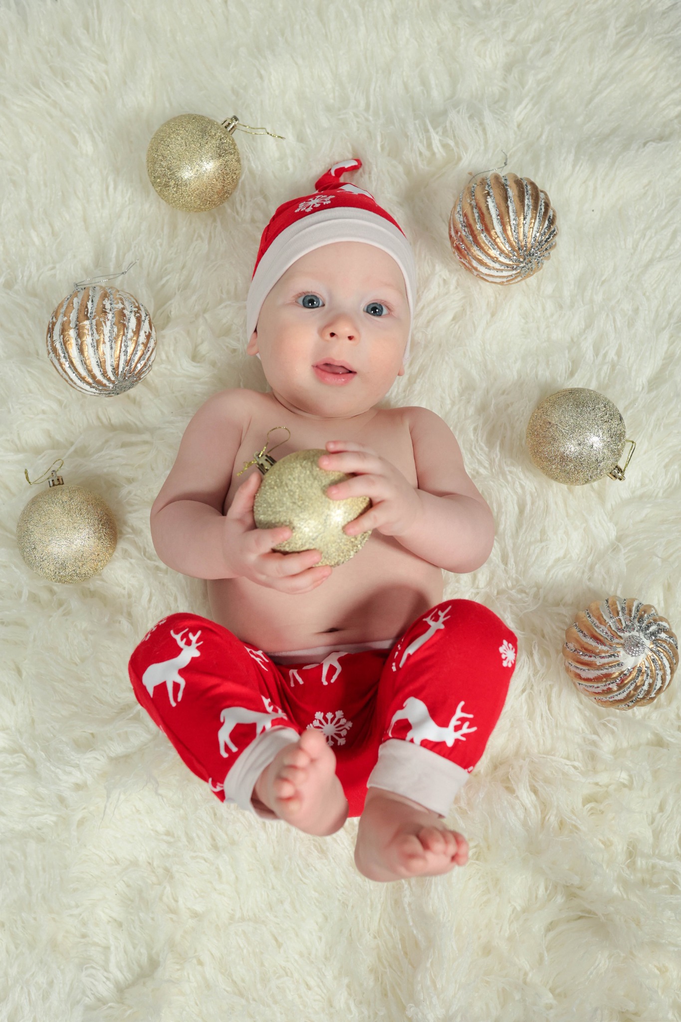 Niemowlę w czerwonej czapce Mikołaja i spodniach z reniferami leży na białym futrzanym dywanie, trzymając złotą bombkę i otoczone innymi ozdobami świątecznymi.