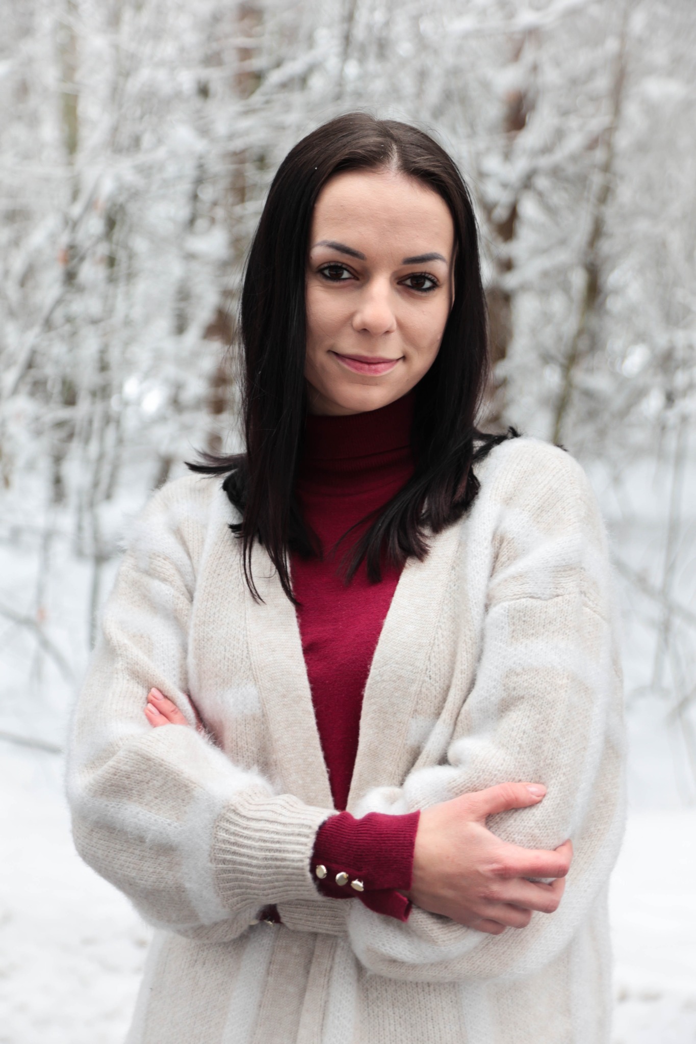 Portret kobiety z ciemnymi włosami, w czerwonym golfie i kremowym kardiganie, na tle zimowego lasu.