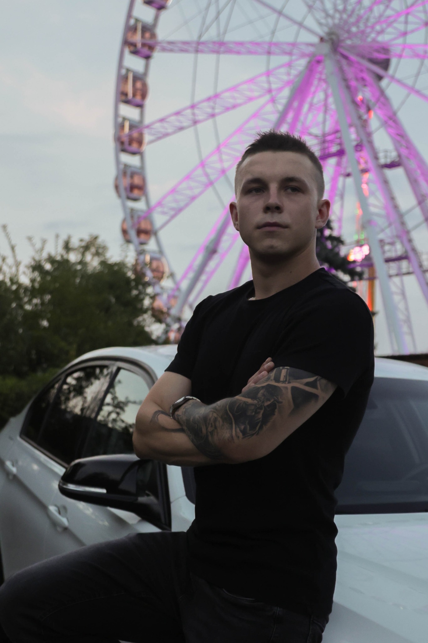 Młody mężczyzna z tatuażami na rękach, oparty o biały samochód, na tle oświetlonego diabelskiego młyna.