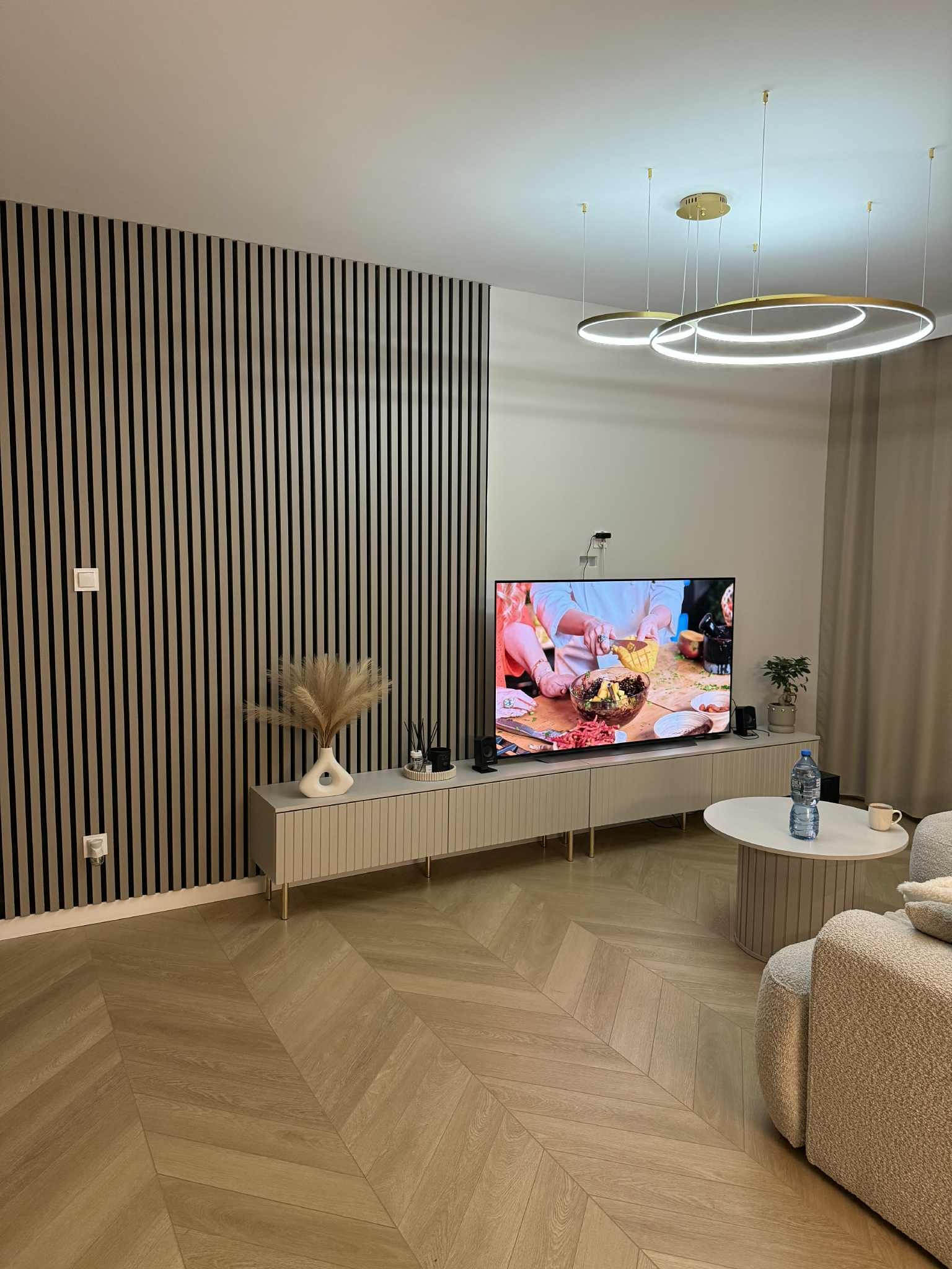 Nowoczesny salon z lamelami na ścianie, parkietem w jodełkę i designerskim oświetleniem. Telewizor wyświetla kulinarny program. Minimalistyczny, jasny wystrój.