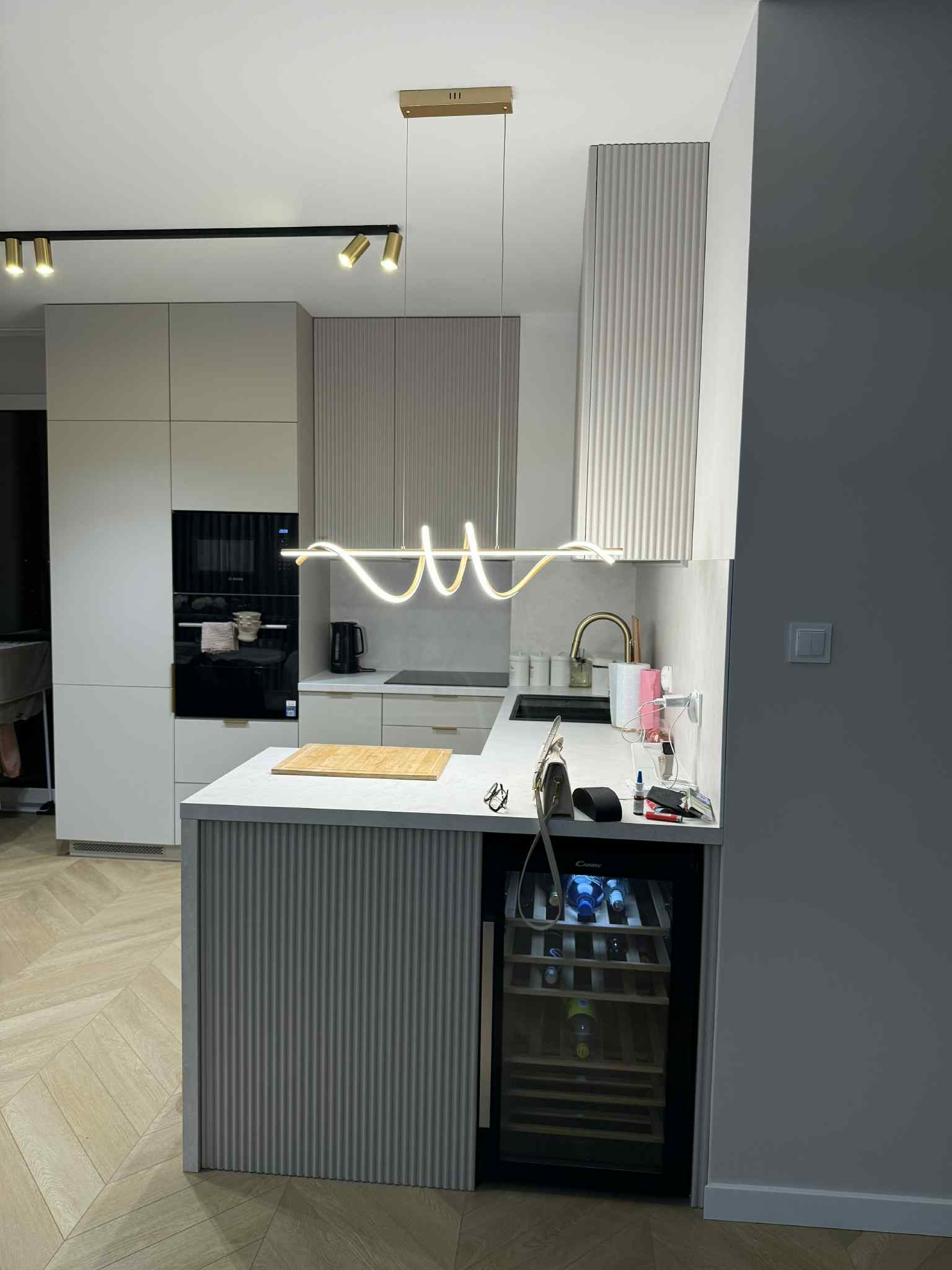 Nowoczesna kuchnia z wyspą, oświetleniem LED i barkiem na wino. Uwagę przykuwają ryflowane fronty szafek i parkiet ułożony w jodełkę.