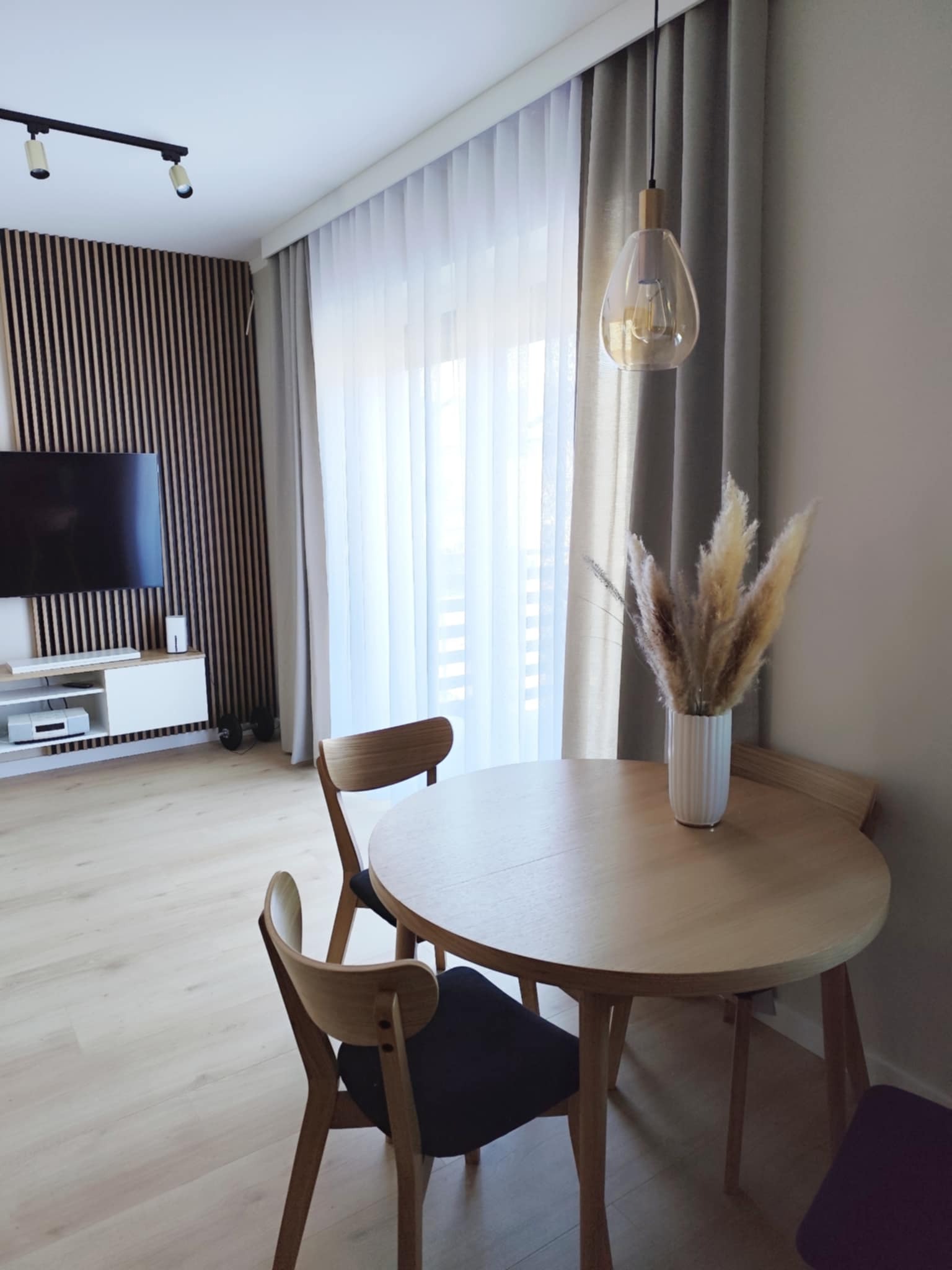 Jasne wnętrze: okrągły stół z drewnianym blatem, krzesła z ciemnymi siedziskami i wazon z trawą pampasową. W tle ściana z drewnianych lameli i okno z zasłonami.