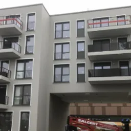 Nowoczesny budynek wielorodzinny z balkonami i szarą elewacją, widoczny podnośnik koszowy firmy Salgert w prześwicie budynku.