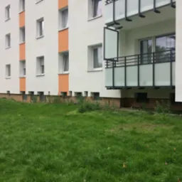 Fragment odnowionego bloku mieszkalnego z balkonem, pomalowanego na biało z pomarańczowymi akcentami, widok z poziomu trawnika.