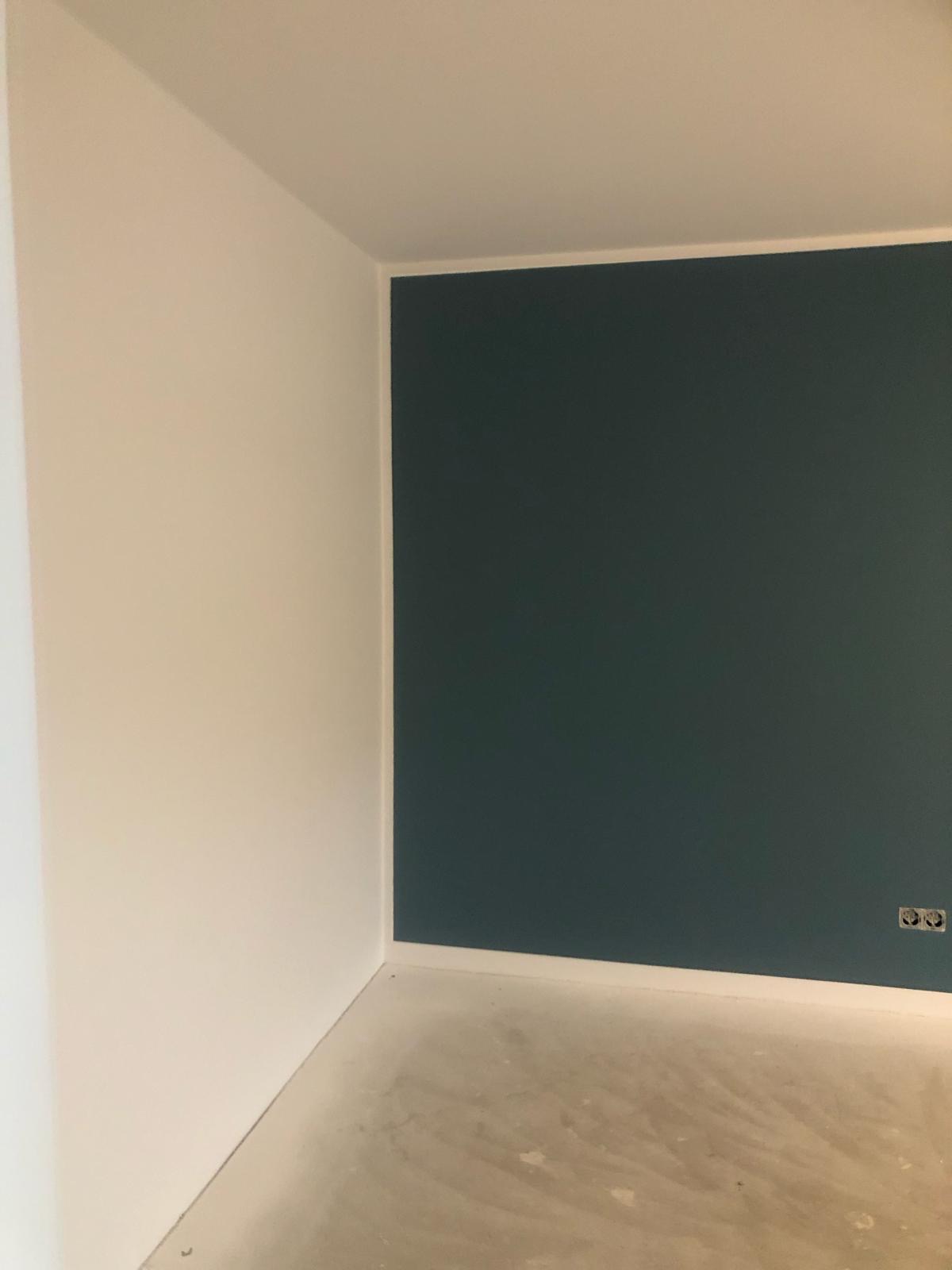 Pomieszczenie po malowaniu ścian: jedna ściana biała, druga pomalowana na ciemny zielony kolor, widoczne gniazdka elektryczne, surowa podłoga przed wykończeniem.