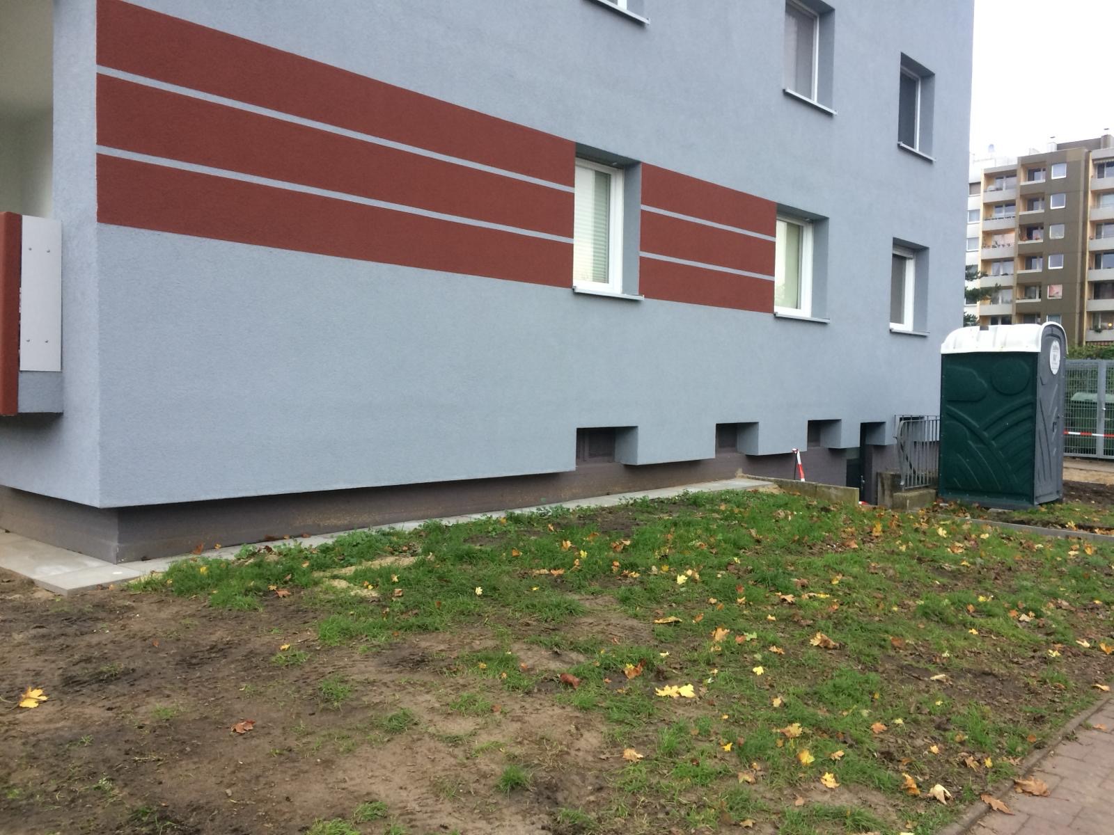 Wykończona elewacja budynku wielorodzinnego w kolorze szarym z poziomymi pasami w odcieniach brązu, widoczna z poziomu gruntu z fragmentem trawnika i przenośną toaletą.