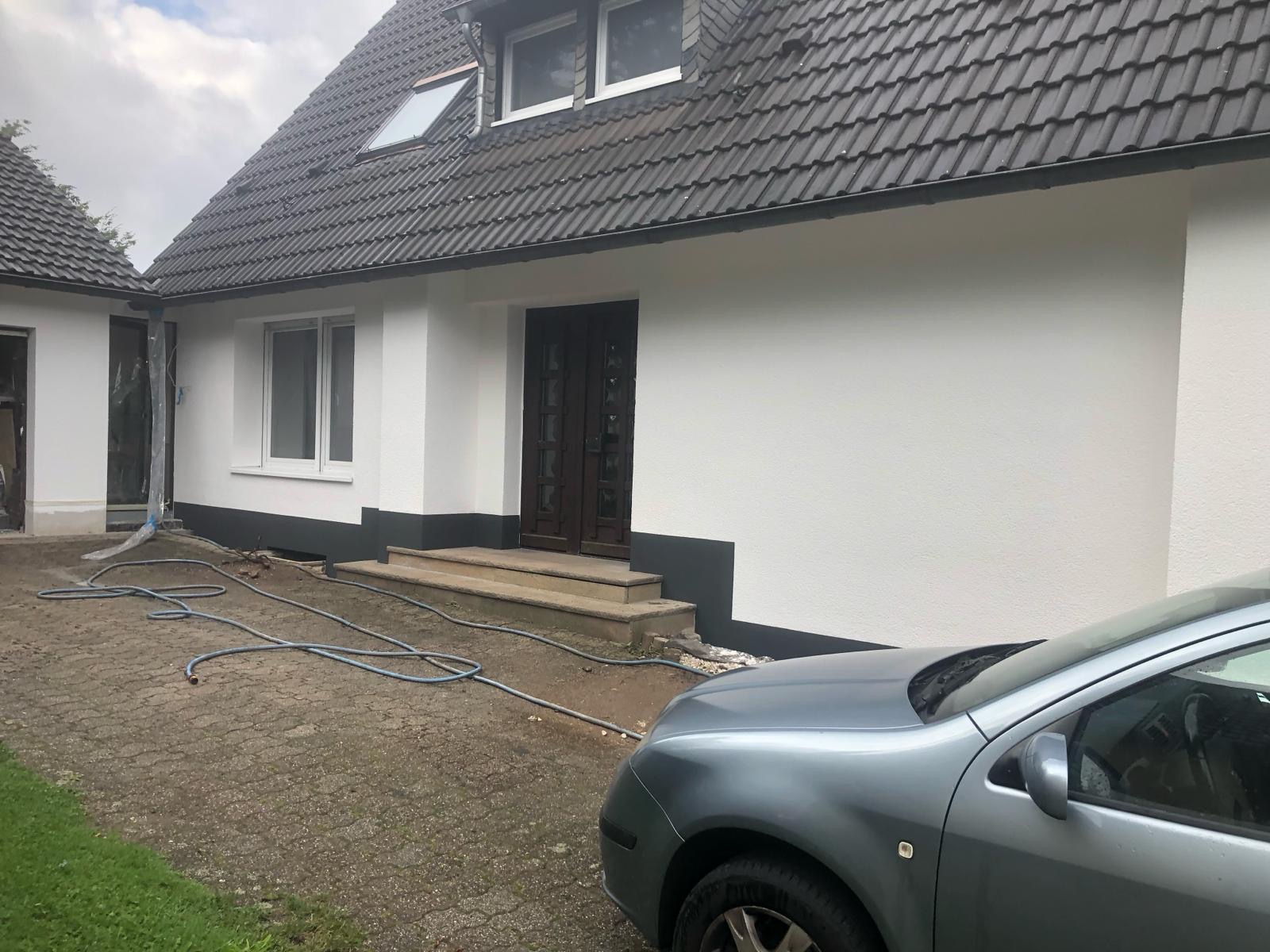 Biała elewacja domu jednorodzinnego z ciemną podmurówką i grafitowym dachem, widoczny fragment samochodu na pierwszym planie oraz zwinięty wąż ogrodowy na brukowanym podjeździe.