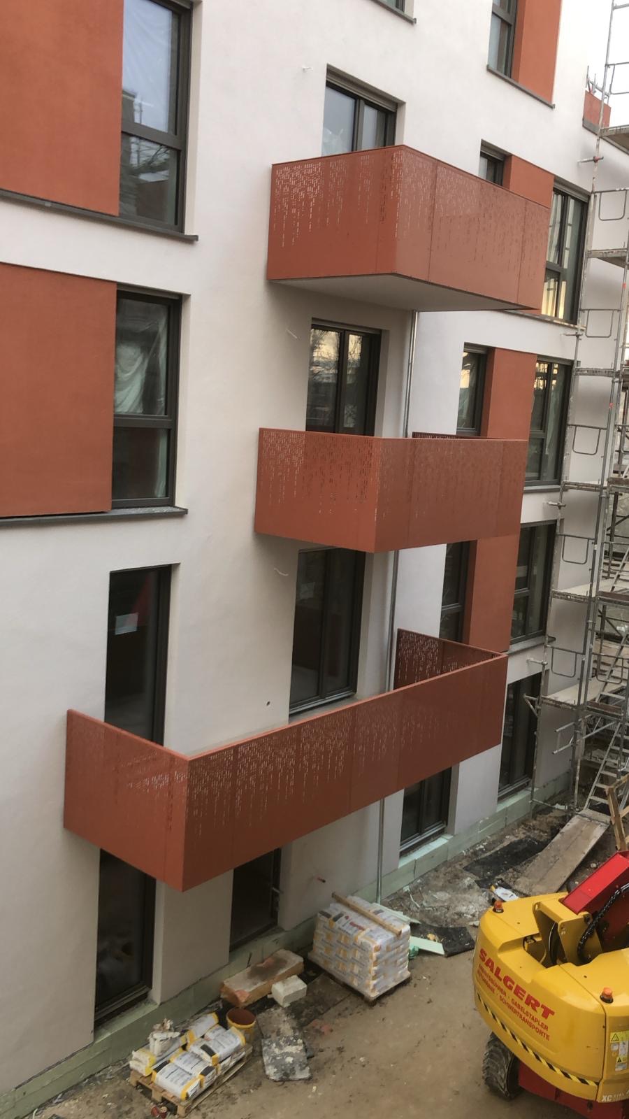 Nowoczesny budynek z elewacją w kolorze białym i pomarańczowym, z ażurowymi balkonami w kolorze pomarańczowym, widok z góry na plac budowy z materiałami budowlanymi i podnośnikiem.