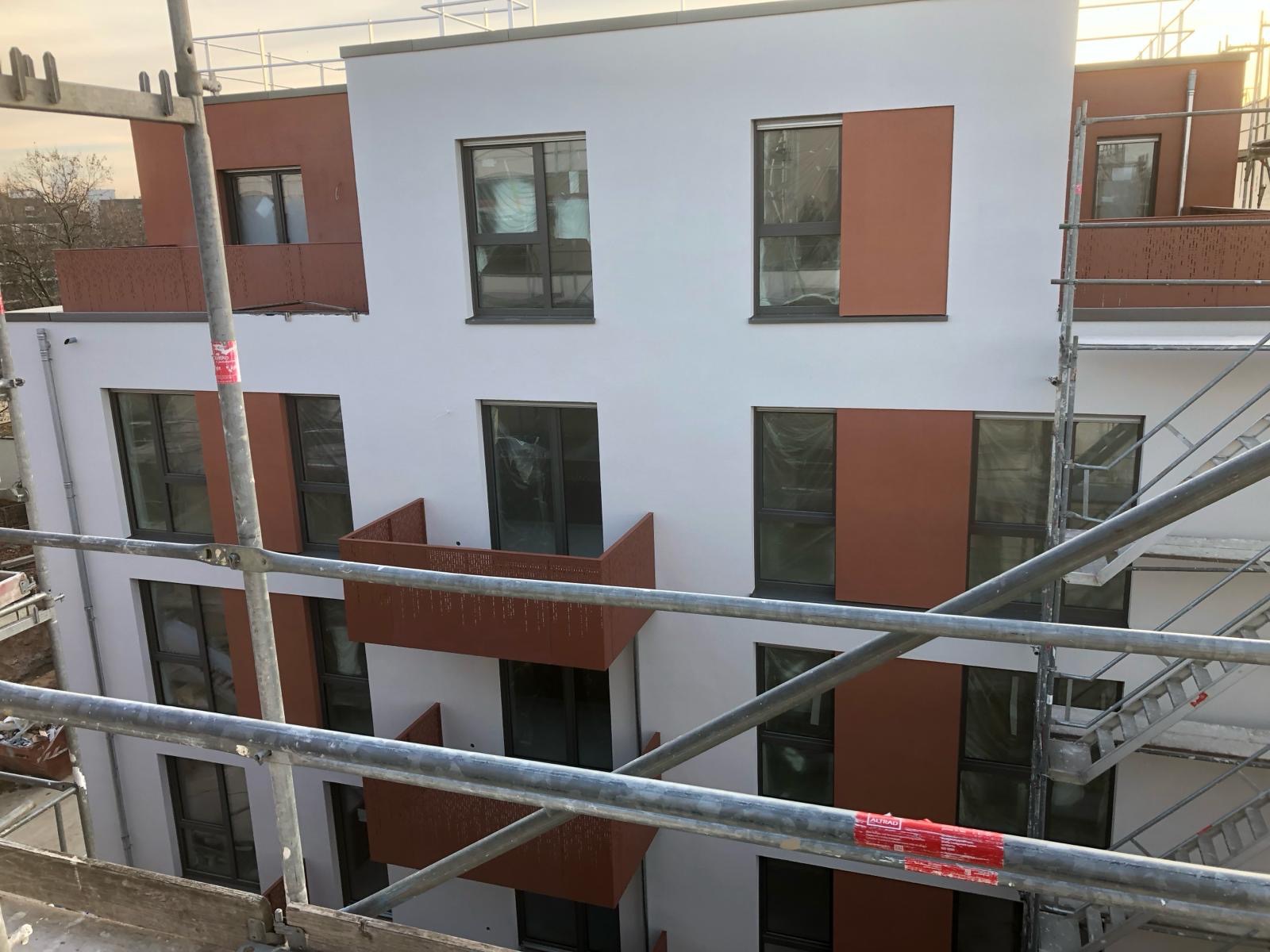 Nowoczesny budynek wielorodzinny z fragmentami elewacji w kolorze terakoty, częściowo zakryty rusztowaniami, okna zabezpieczone folią.