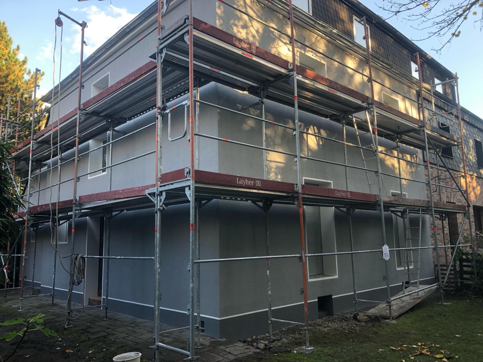 Świeżo otynkowana elewacja budynku w szarym kolorze, zabezpieczona rusztowaniem Layher, widoczne detale wykończenia wokół okien i u podstawy ściany.