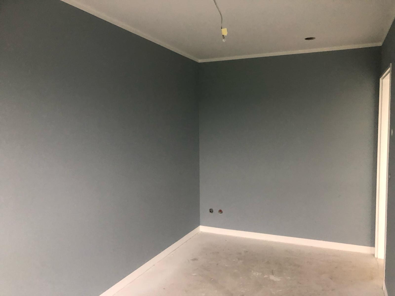 Wnętrze pomalowane na szaro z białymi listwami przypodłogowymi i sufitowymi, widoczne gniazdka elektryczne i wystające kable z sufitu, surowa podłoga przed wykończeniem.