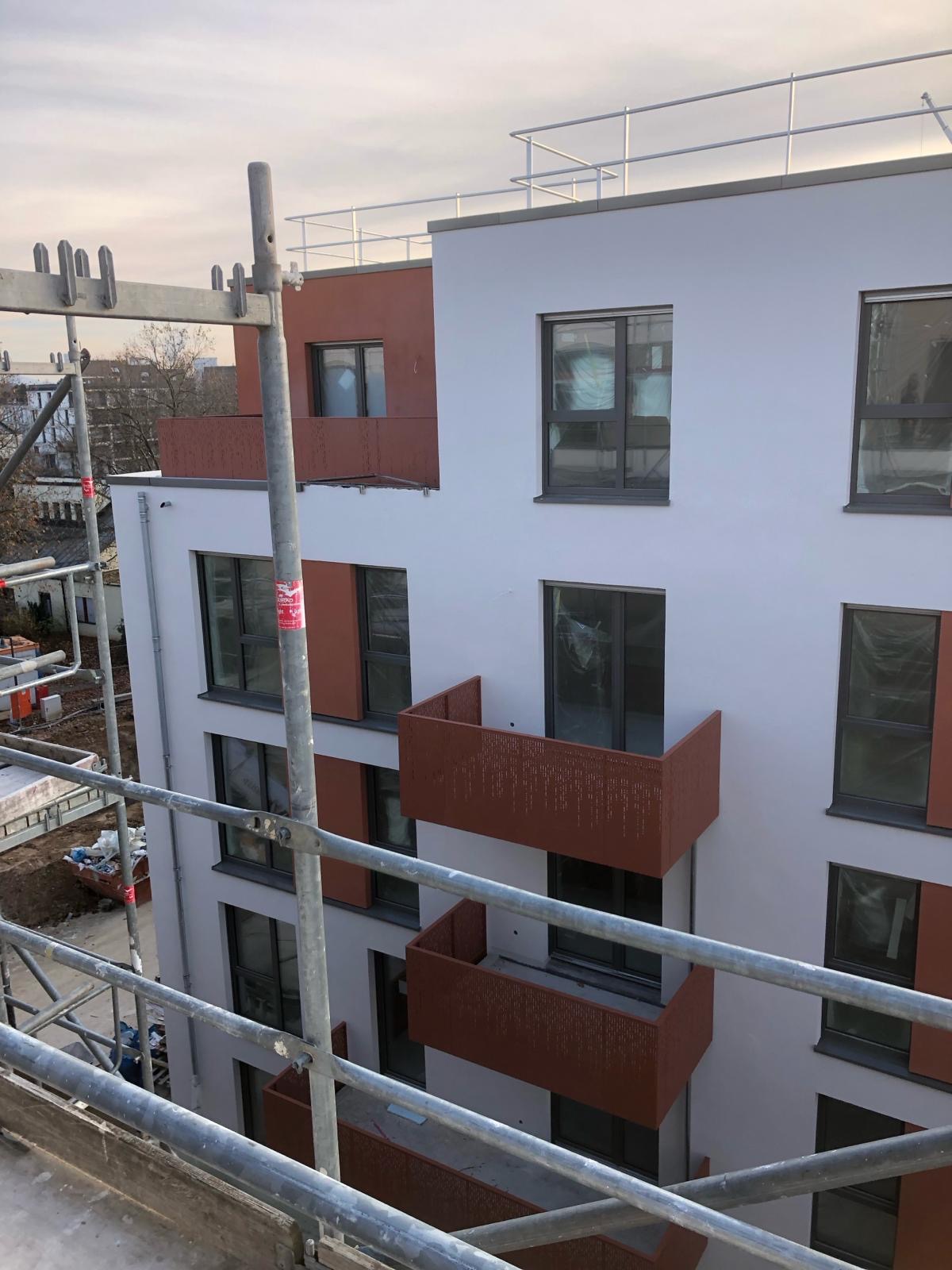 Wielopiętrowy budynek z balkonami o nowoczesnej elewacji w kolorze białym i brązowym, częściowo zasłonięty rusztowaniem.