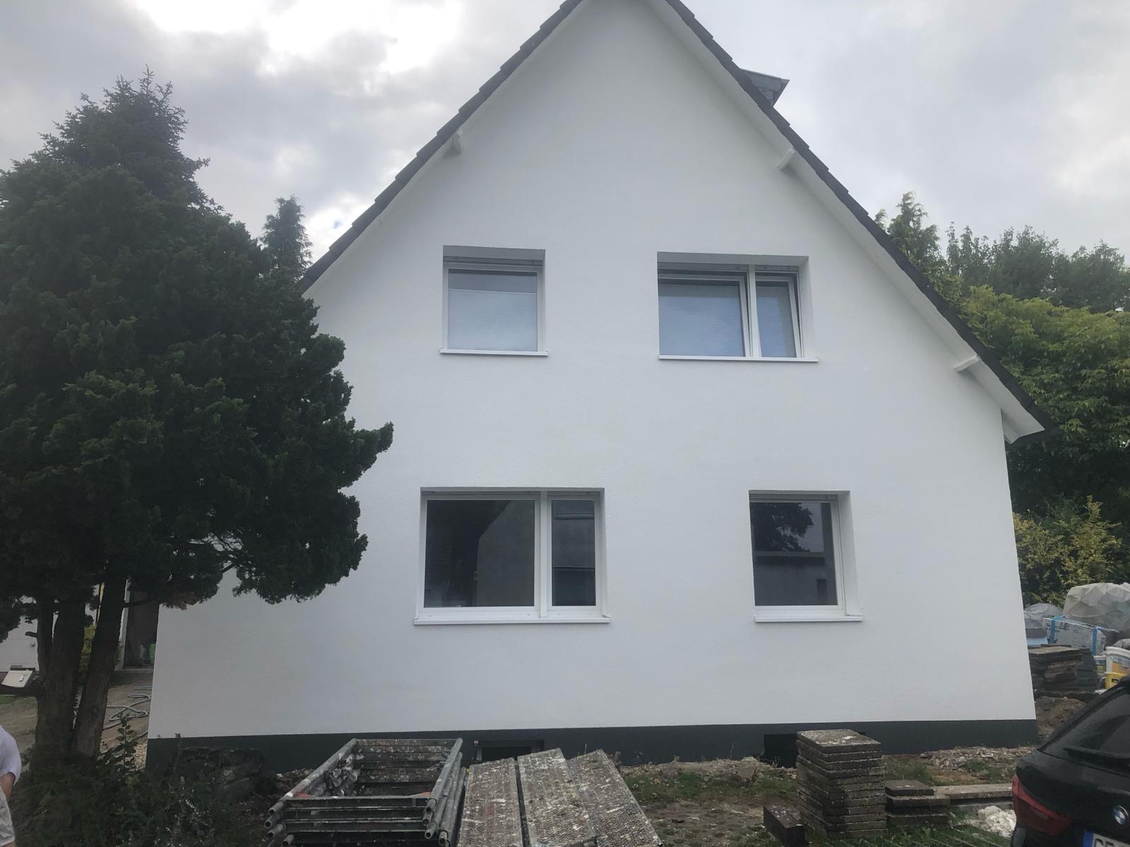 Dwupiętrowy dom z białymi ścianami po renowacji, widoczne rusztowania i drzewo iglaste po lewej stronie, pochmurne niebo w tle.