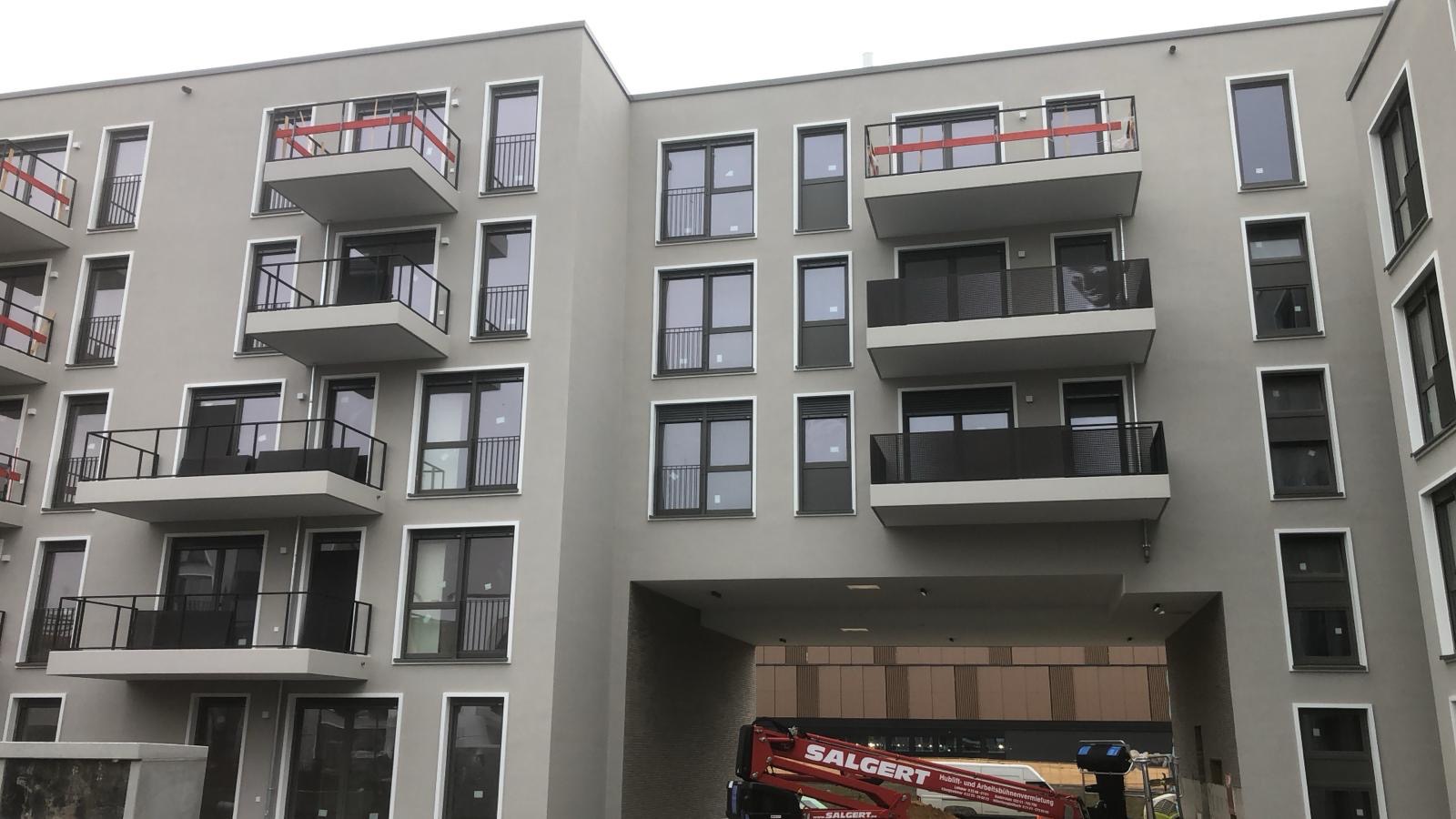 Nowoczesny budynek wielorodzinny z balkonami i szarą elewacją, widoczny podnośnik koszowy firmy Salgert w prześwicie budynku.