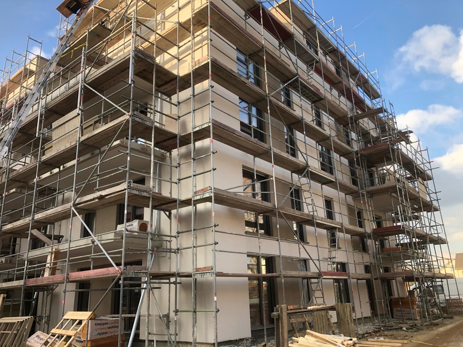 Wielopiętrowy budynek w trakcie prac elewacyjnych, otoczony rusztowaniem, z widocznymi fragmentami ocieplenia i świeżo położonego tynku w kolorze jasnego beżu, na dole leżą materiały budowlane.